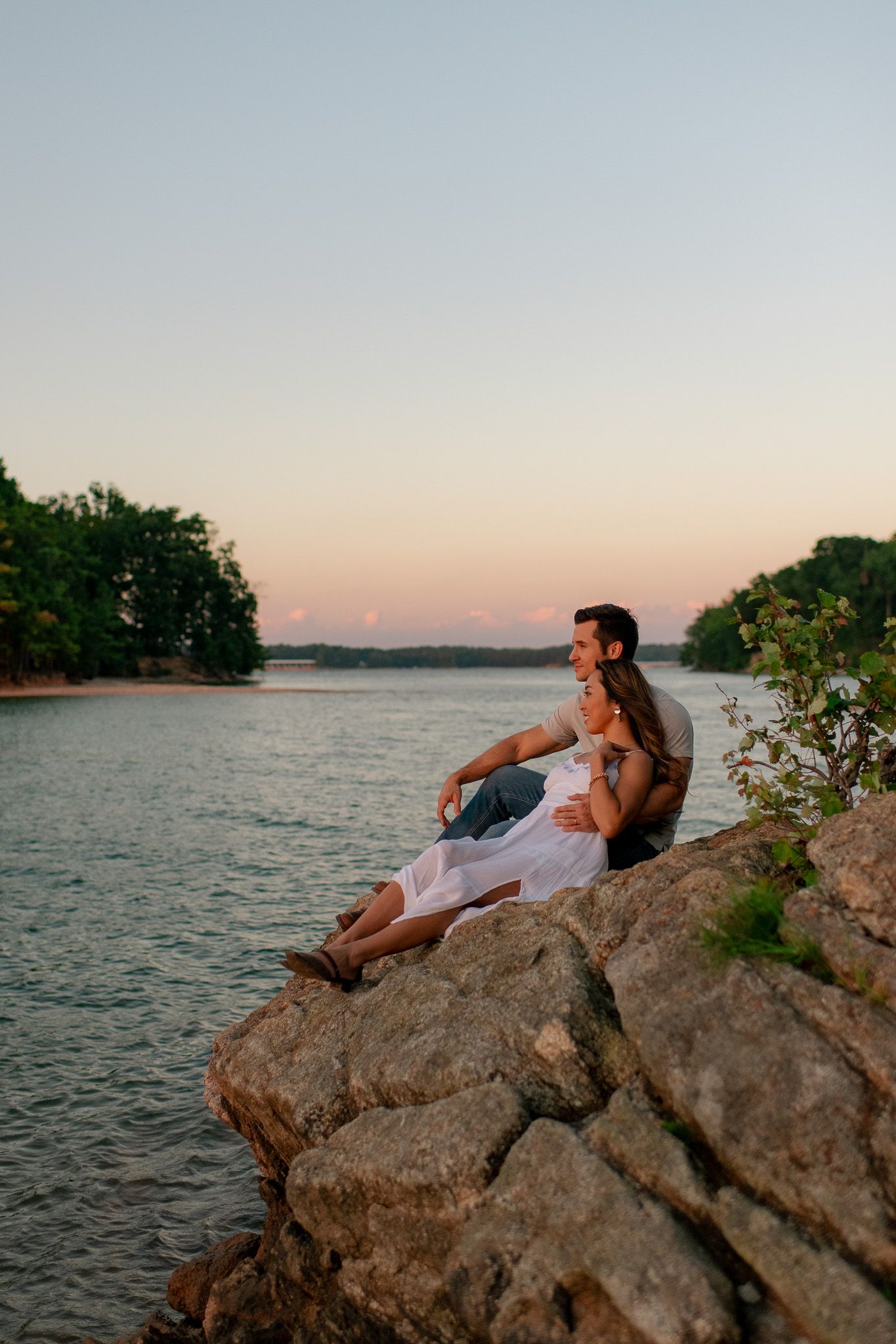 A couple sits together on rocky cliff overlooking a serene lake at sunset, surrounded by tree-lined shores.