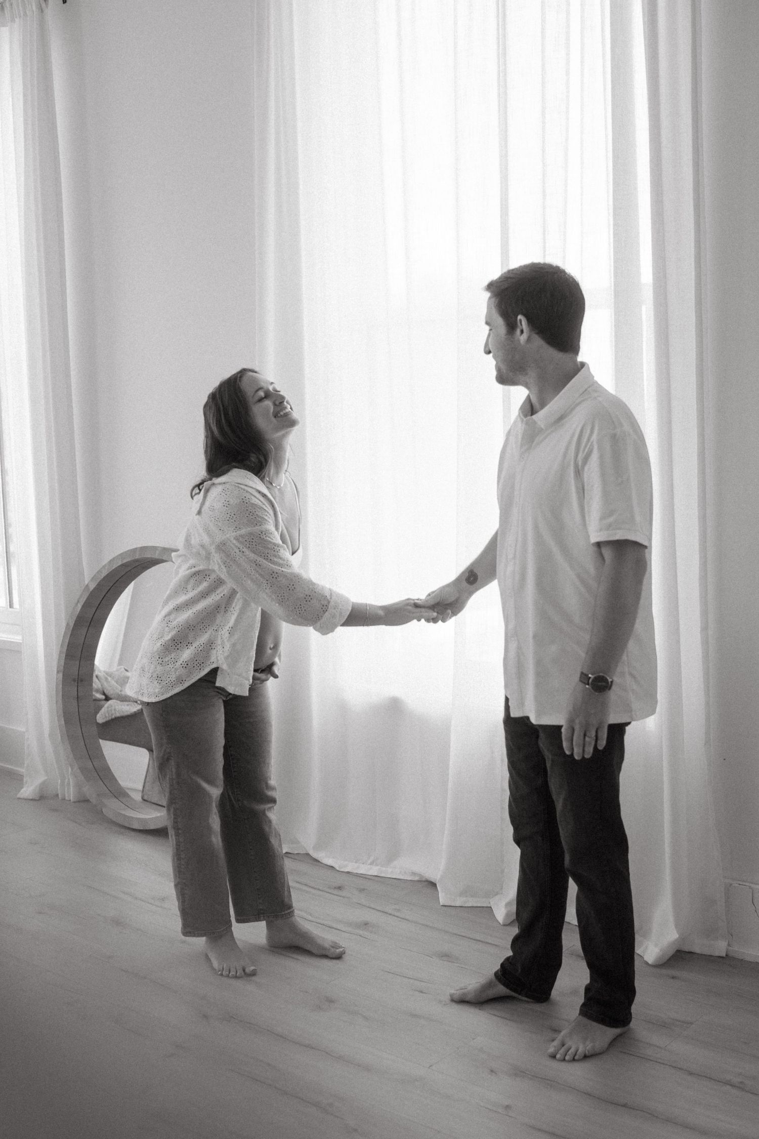Black and white maternity photos showing playful couple interaction by window.
