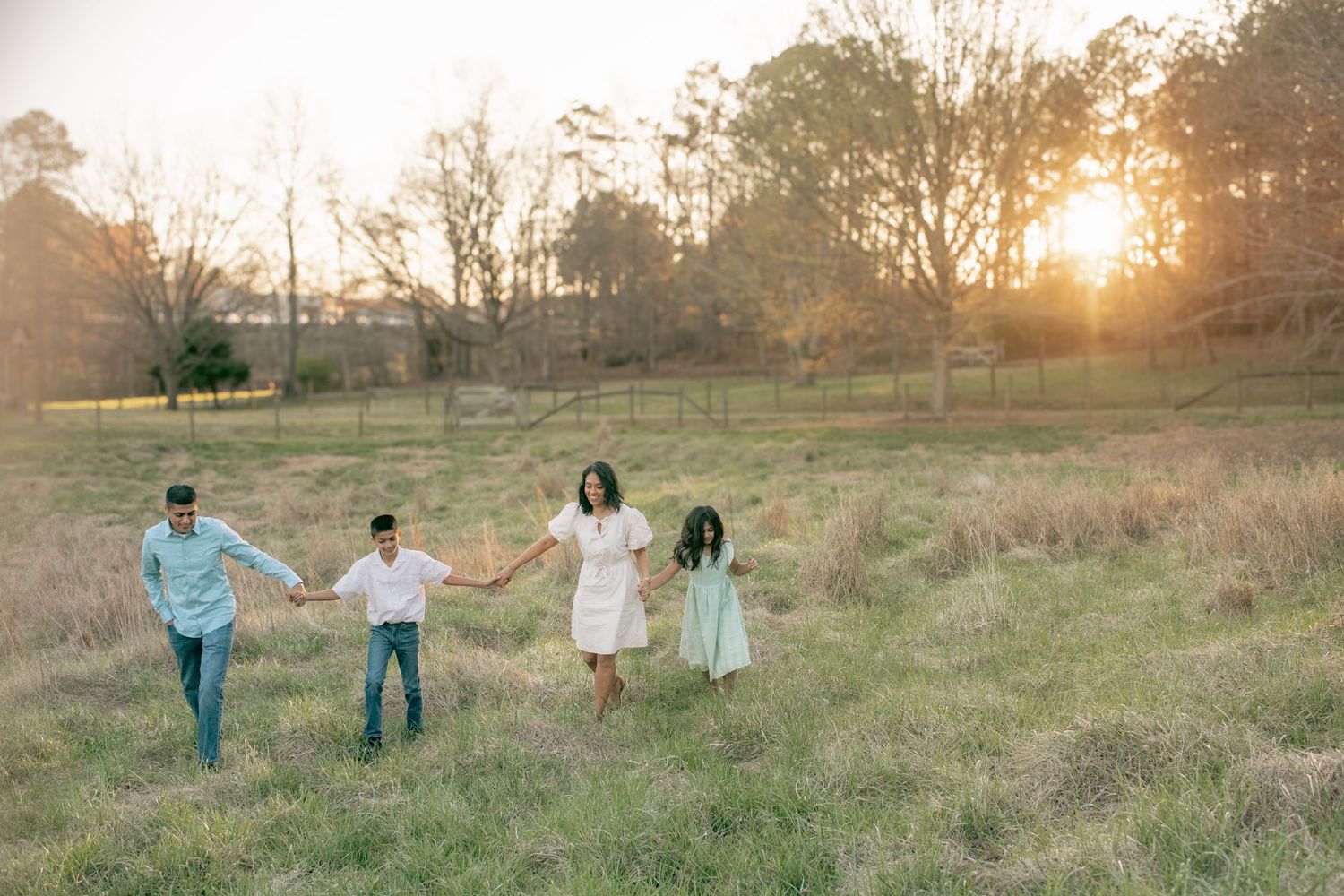 A group holds hands walking through a sun-drenched field at sunset with tall grasses and trees in the background.