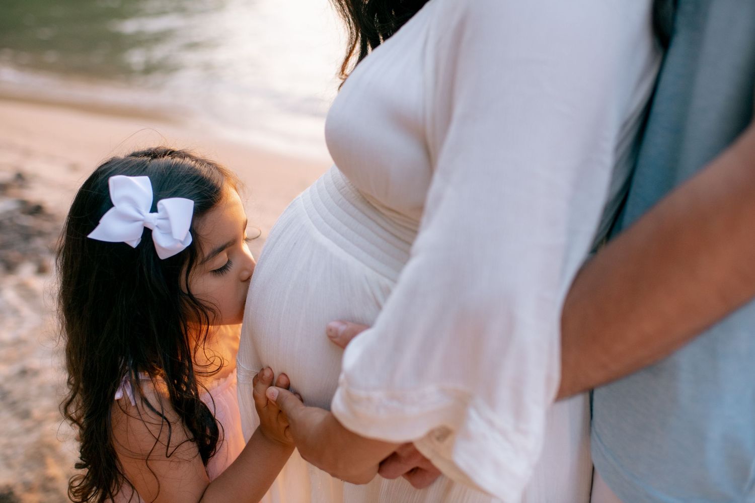 A young child with a white bow in their dark hair looks up at a pregnant woman in a white dress on a beach.