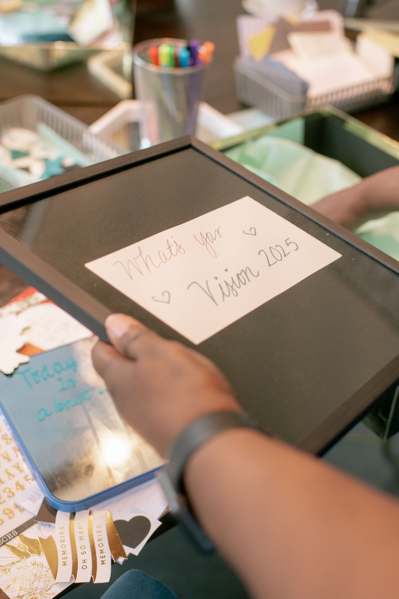 Hand writing on a black digital tablet or device with colorful items visible in the background.