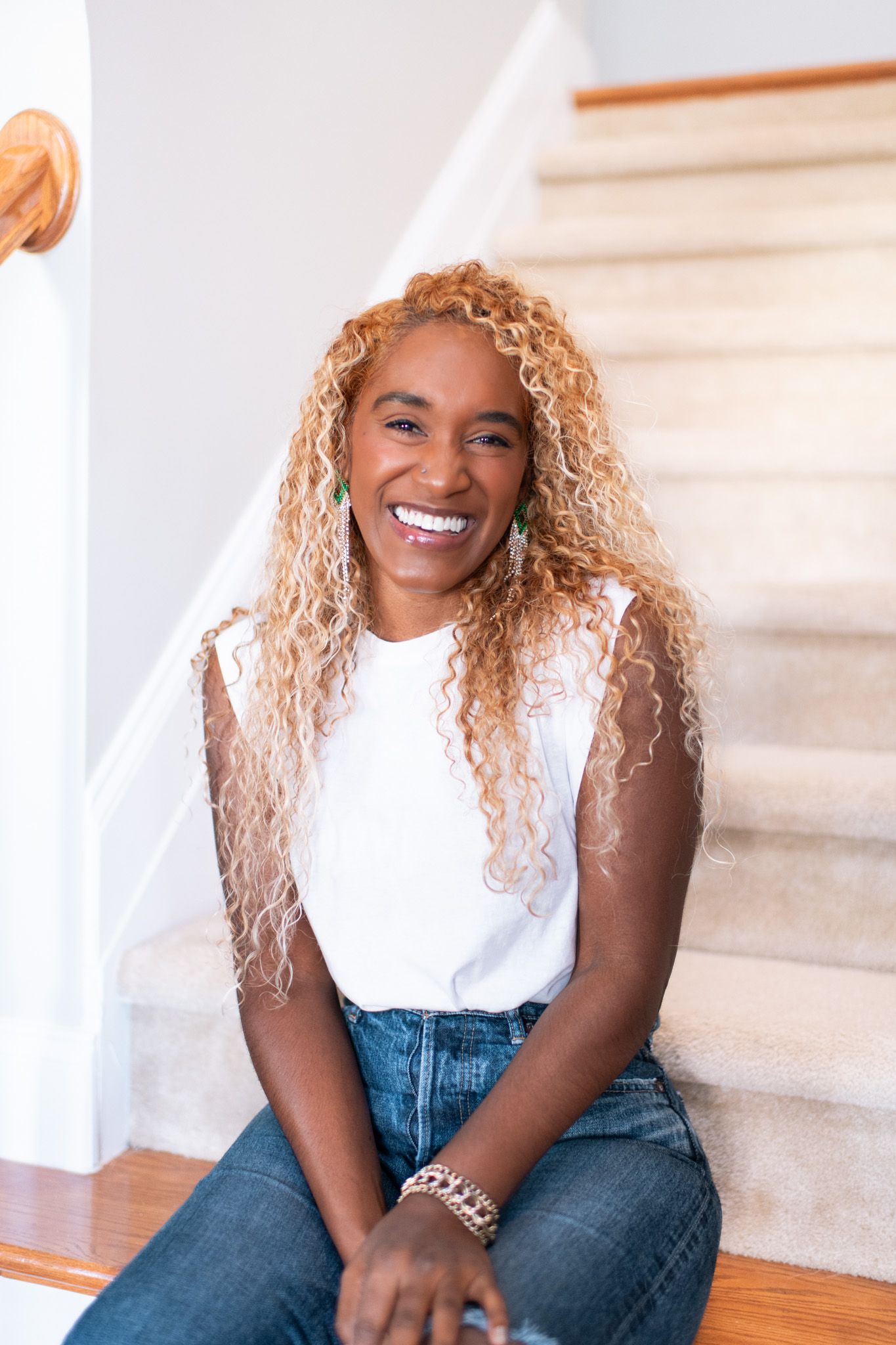 Smiling woman with blonde curly hair sitting on stairs wearing white top and jeans.