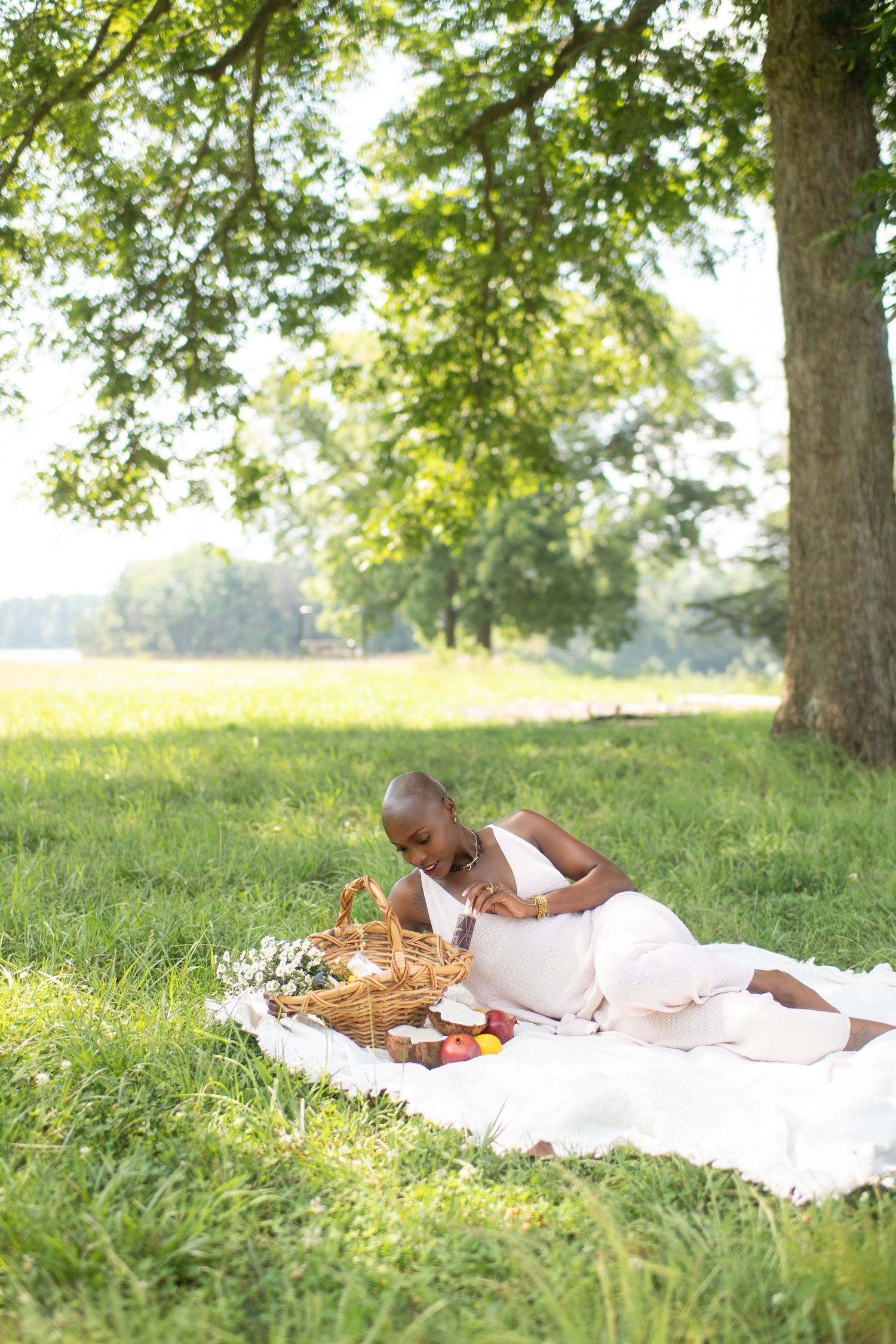Pregnant woman in white dress enjoying a picnic on a blanket under a large tree in a sunny green park.