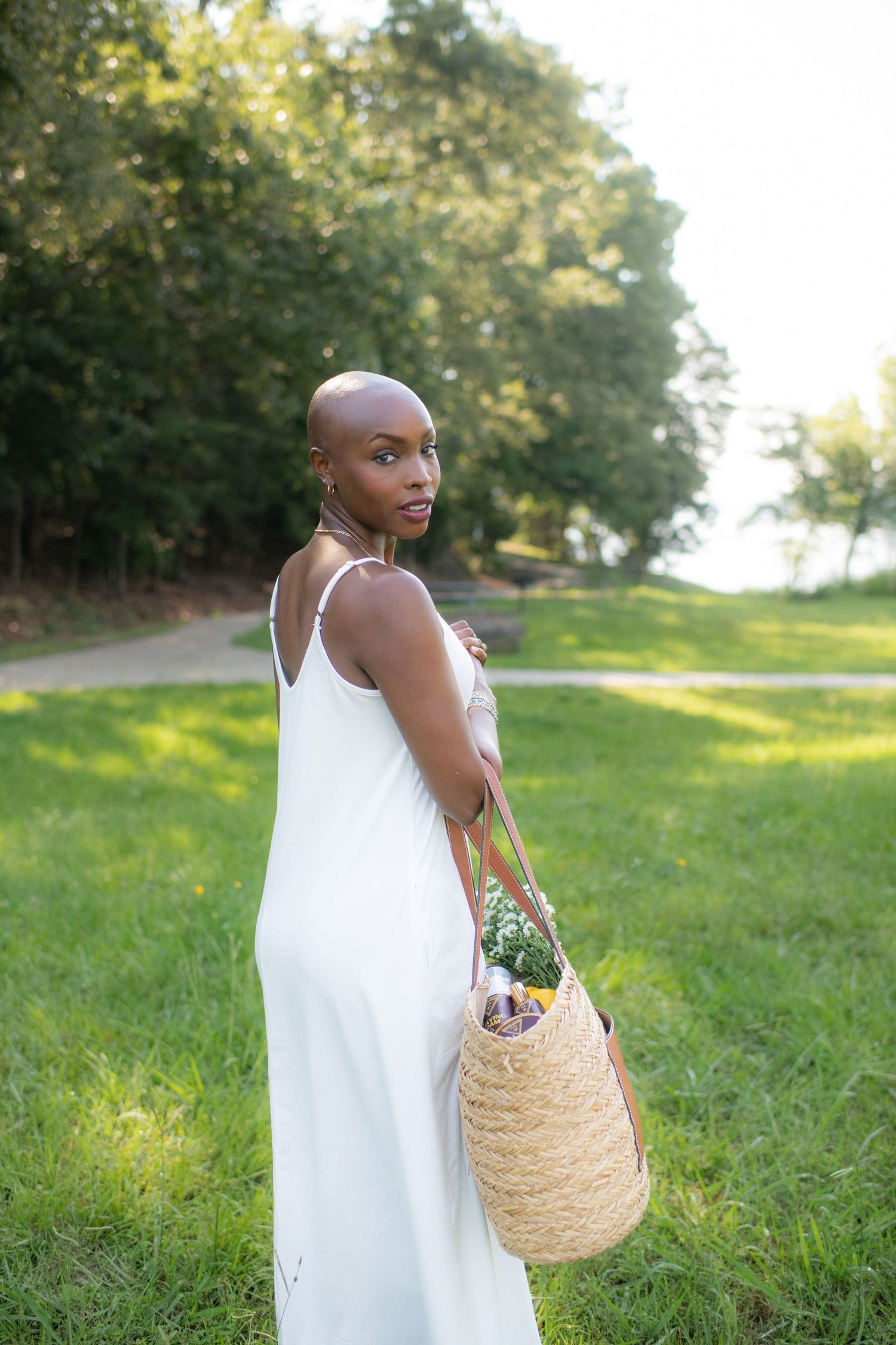 A bald Black woman in a white sundress holds a wicker bag with flowers in a lush green park.