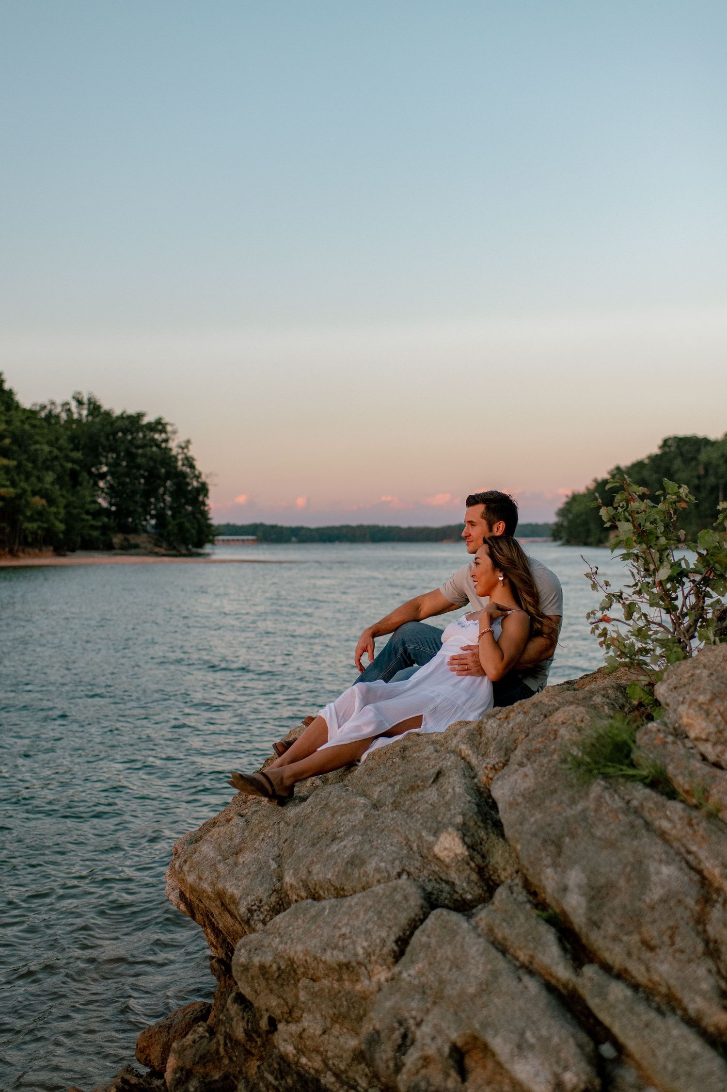 Couple relaxing on rocky outcrop overlooking serene lake at sunset.