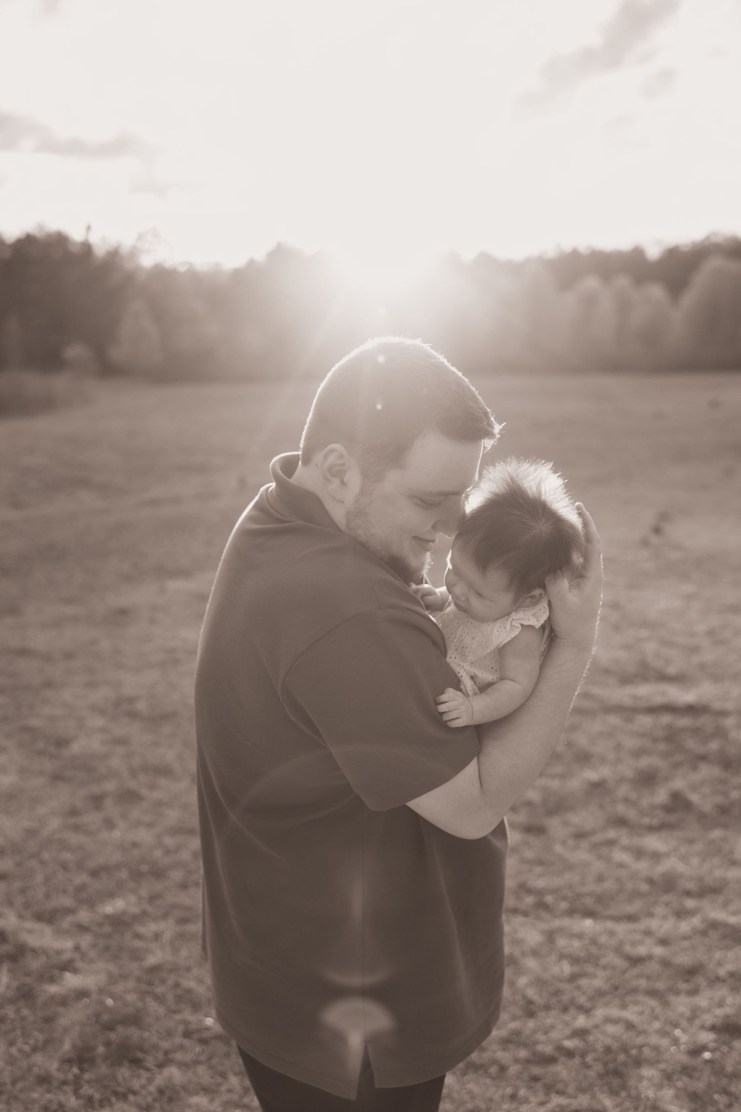 A tender moment captured in sepia tones as a person in a dark sweater embraces a small baby in an outdoor field setting.