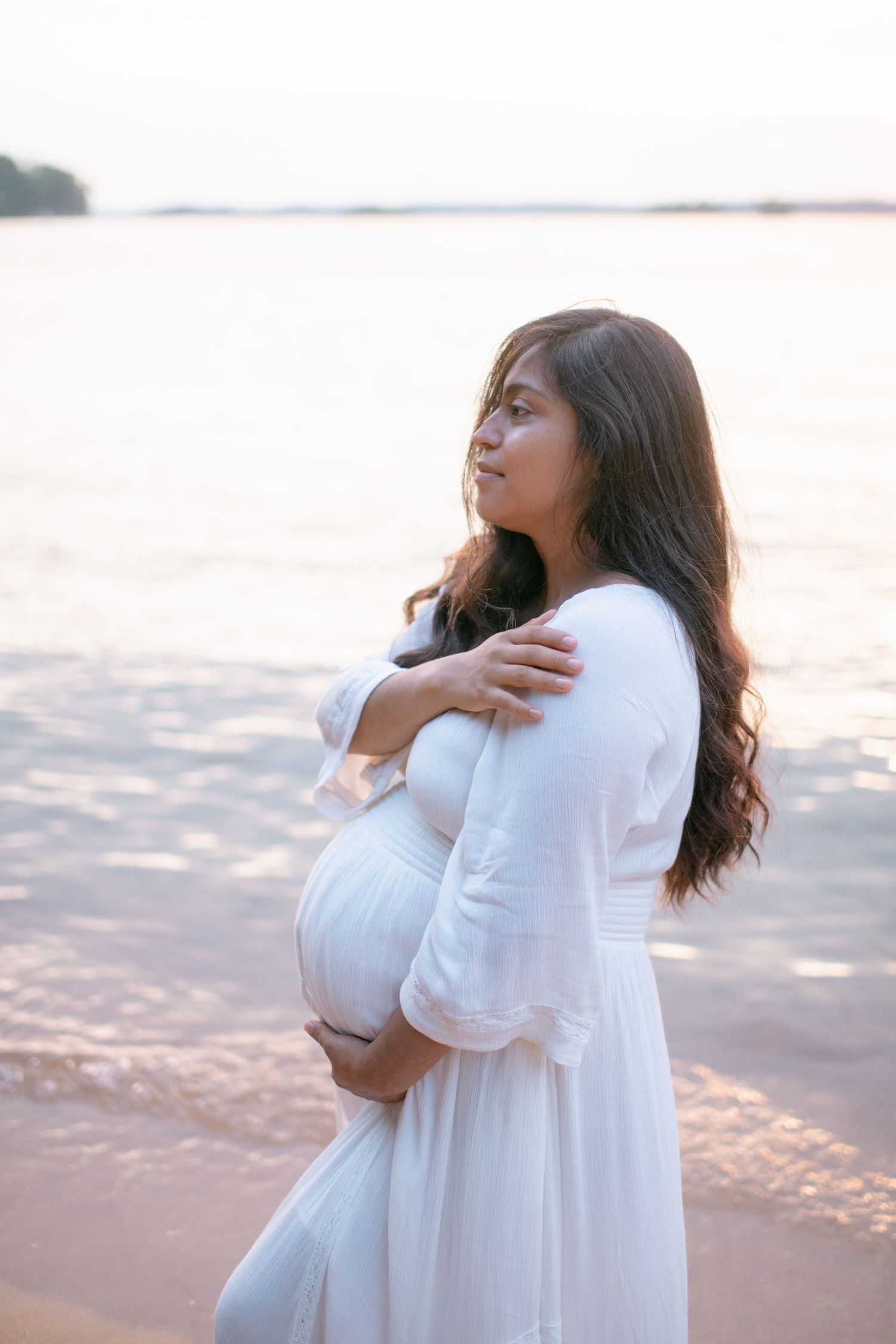 A woman in flowing white dress stands contemplatively at the beach during sunset.