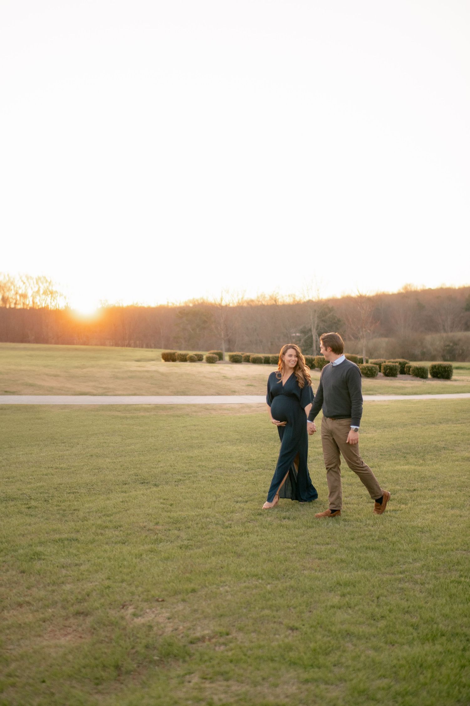 Couple walking together on grassy field at sunset with golden light and trees in background.