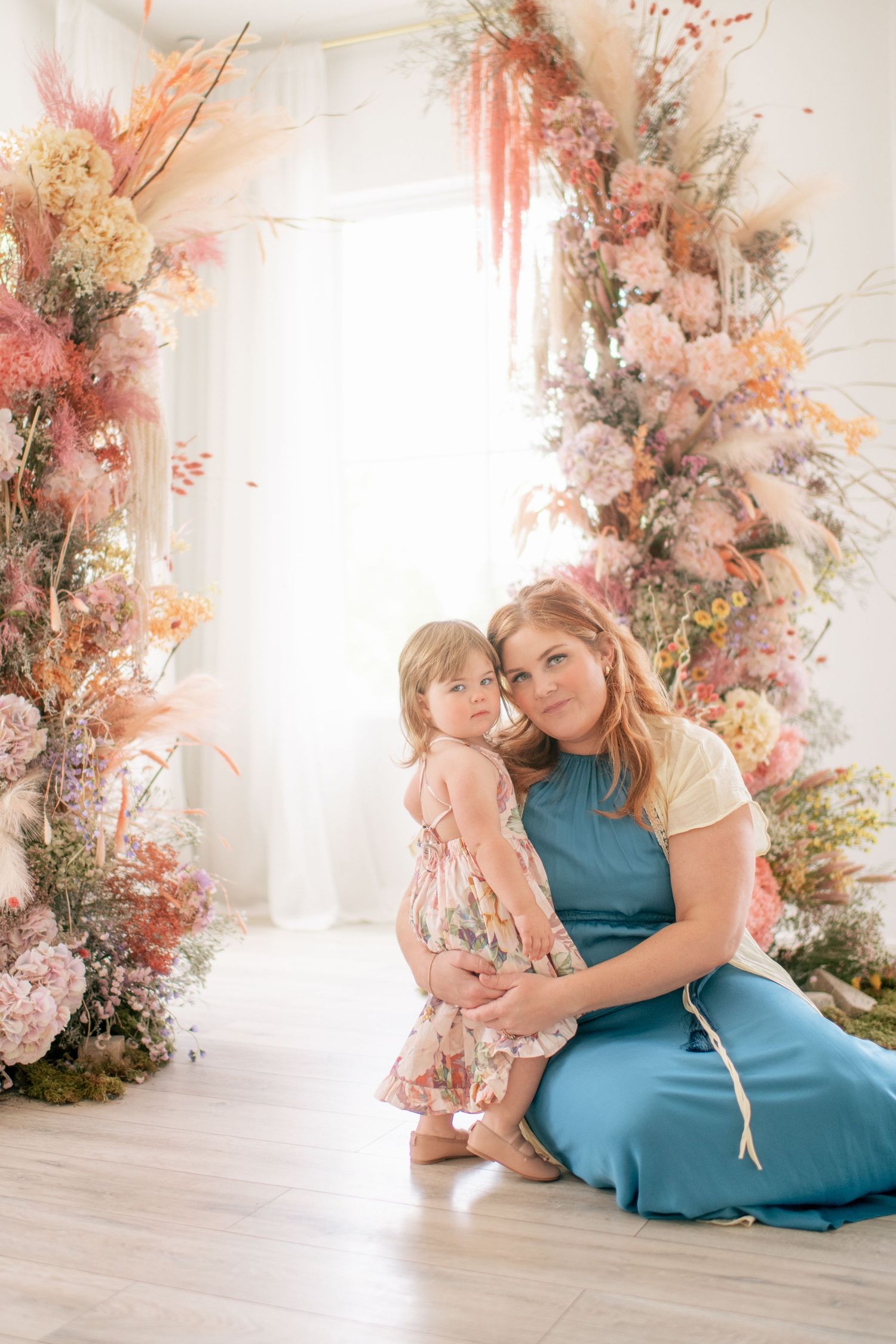 Sweet spring scene with two figures seated beside a beautiful floral arch with pink and peach blooms in a bright studio setting.