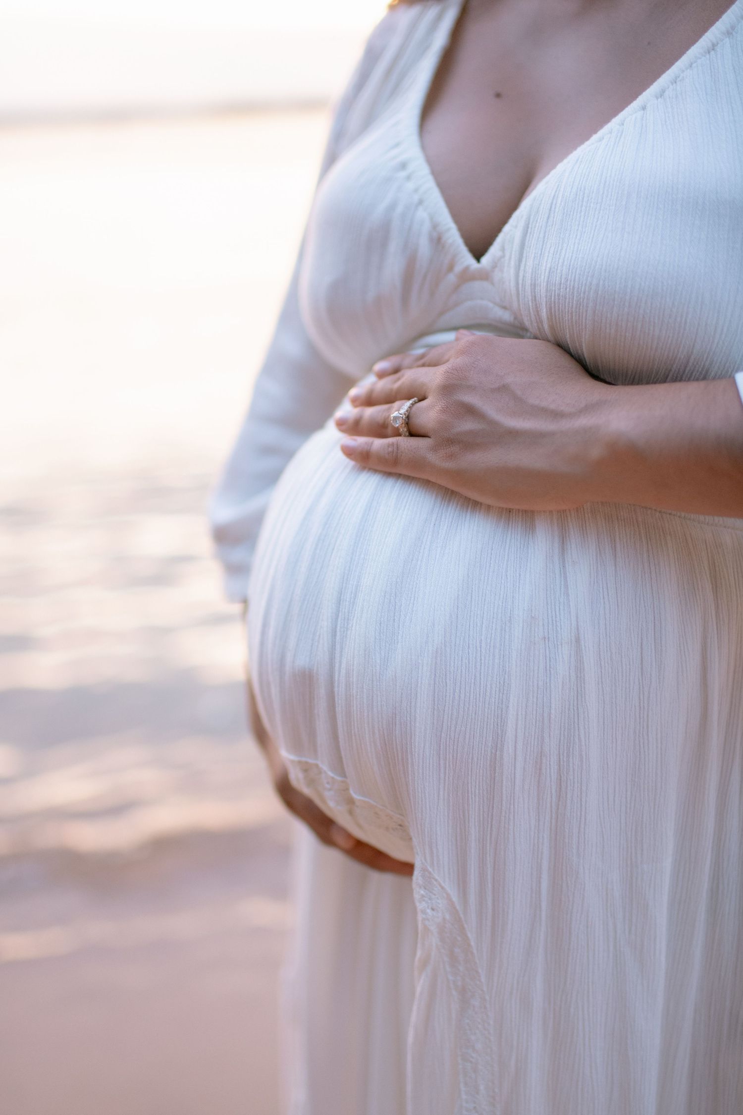 A pregnant woman in a flowing white dress cradles her belly against a soft sunset background at the beach.