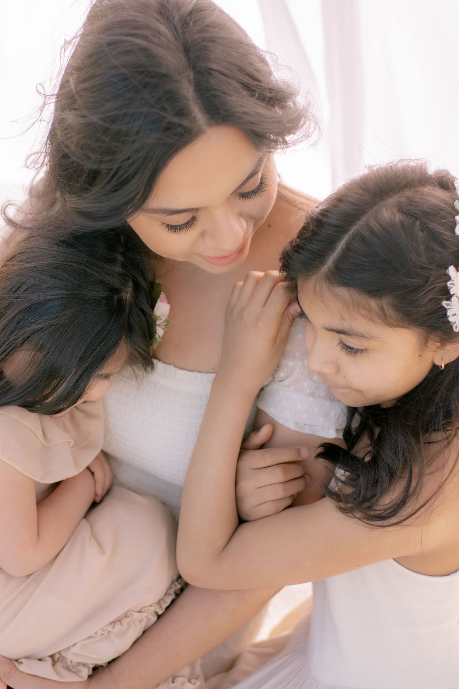 A tender moment captured between a mother and her two daughters in soft lighting and delicate white and blush dresses.