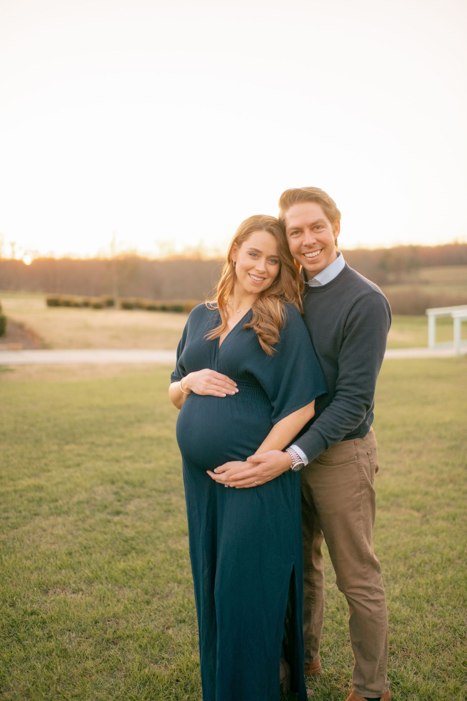 Expectant couple embraces during sunset maternity photo session in grassy field with navy blue dress.