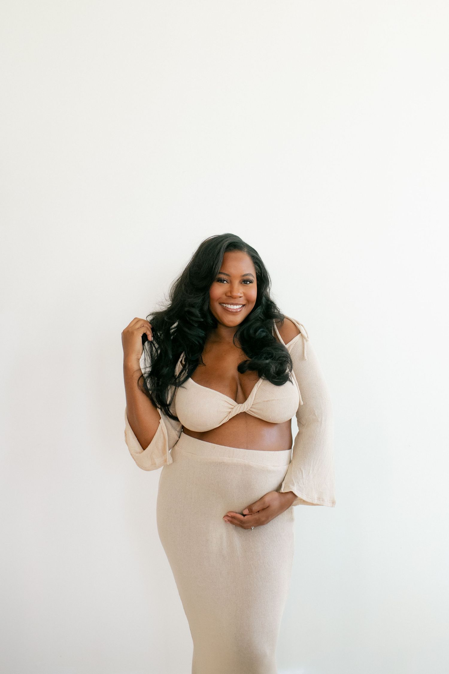 Joyful maternity portrait against white wall in cream-colored two-piece outfit.