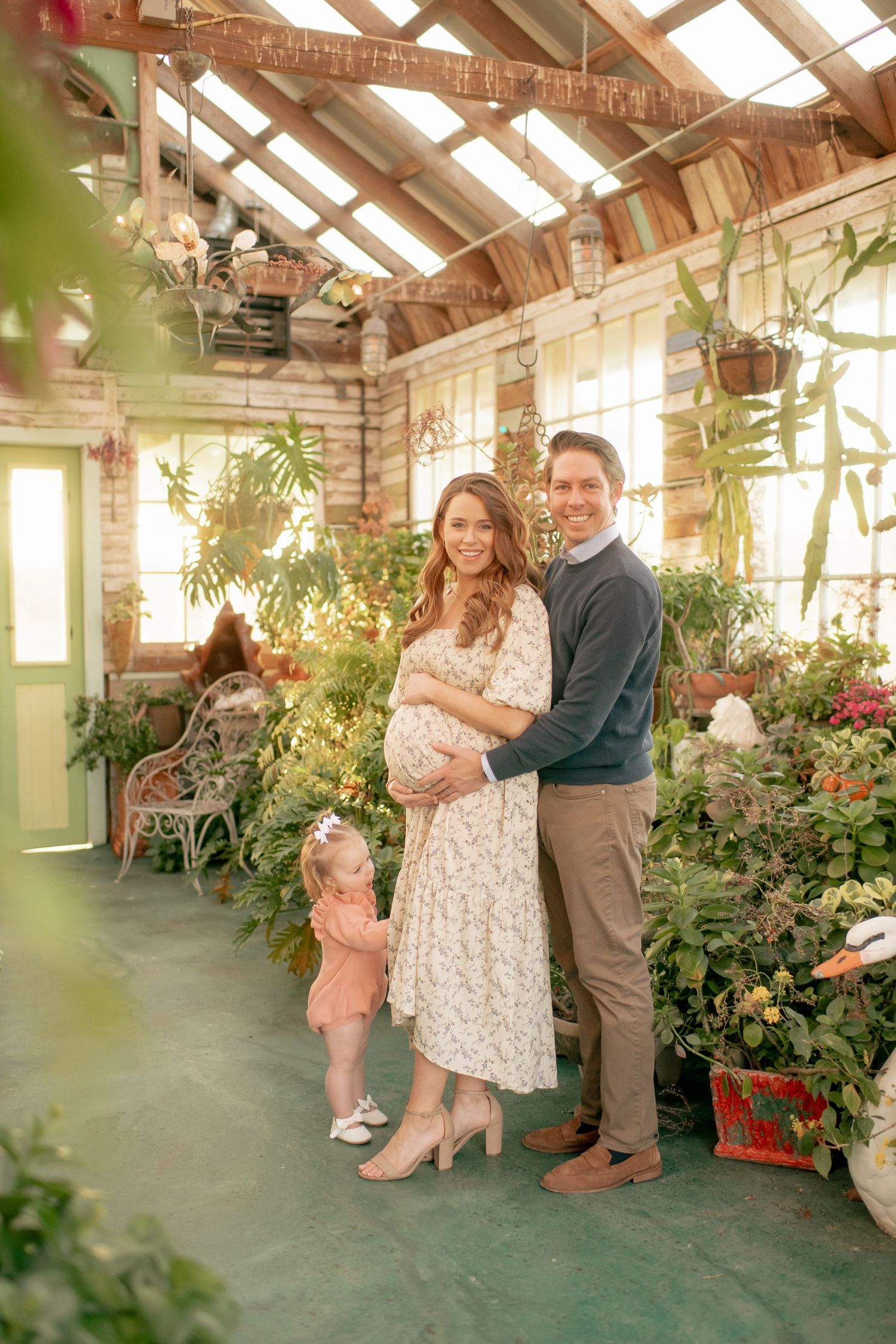 A sun-filled greenhouse setting with hanging plants and flowers creates a warm atmosphere for a family portrait.