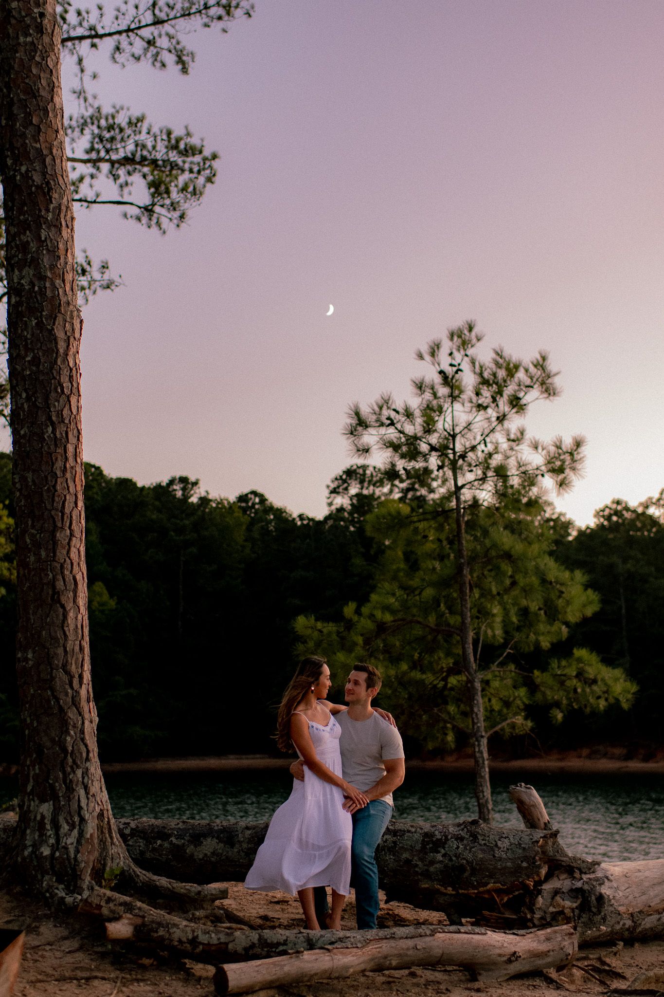Outdoor sunset photoshoot with a pair sitting on fallen tree trunks against a wooded landscape.