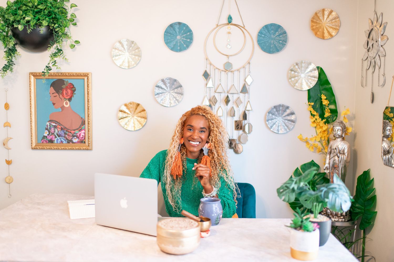 A person in green works on a laptop at a desk decorated with plants and wall art in a boho-style workspace.