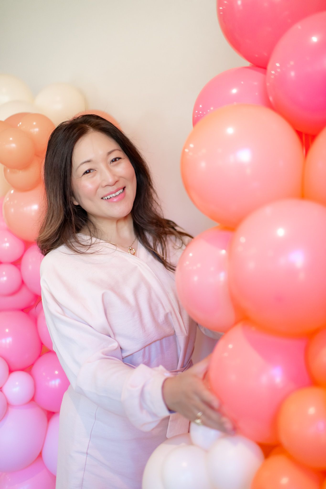 A person in white poses playfully against a backdrop of pink and peach colored balloons at a photo shoot.