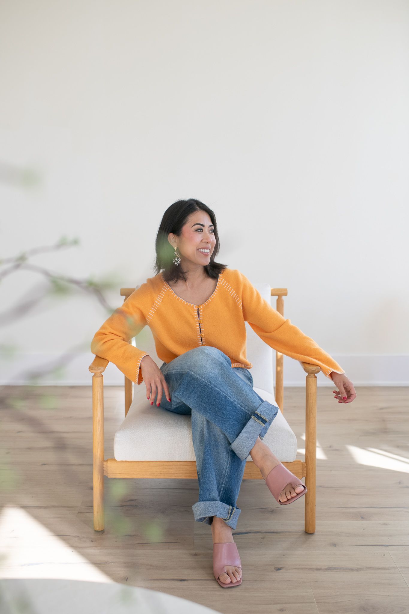 Person in orange sweater and jeans relaxes in wooden chair with green statement earrings.
