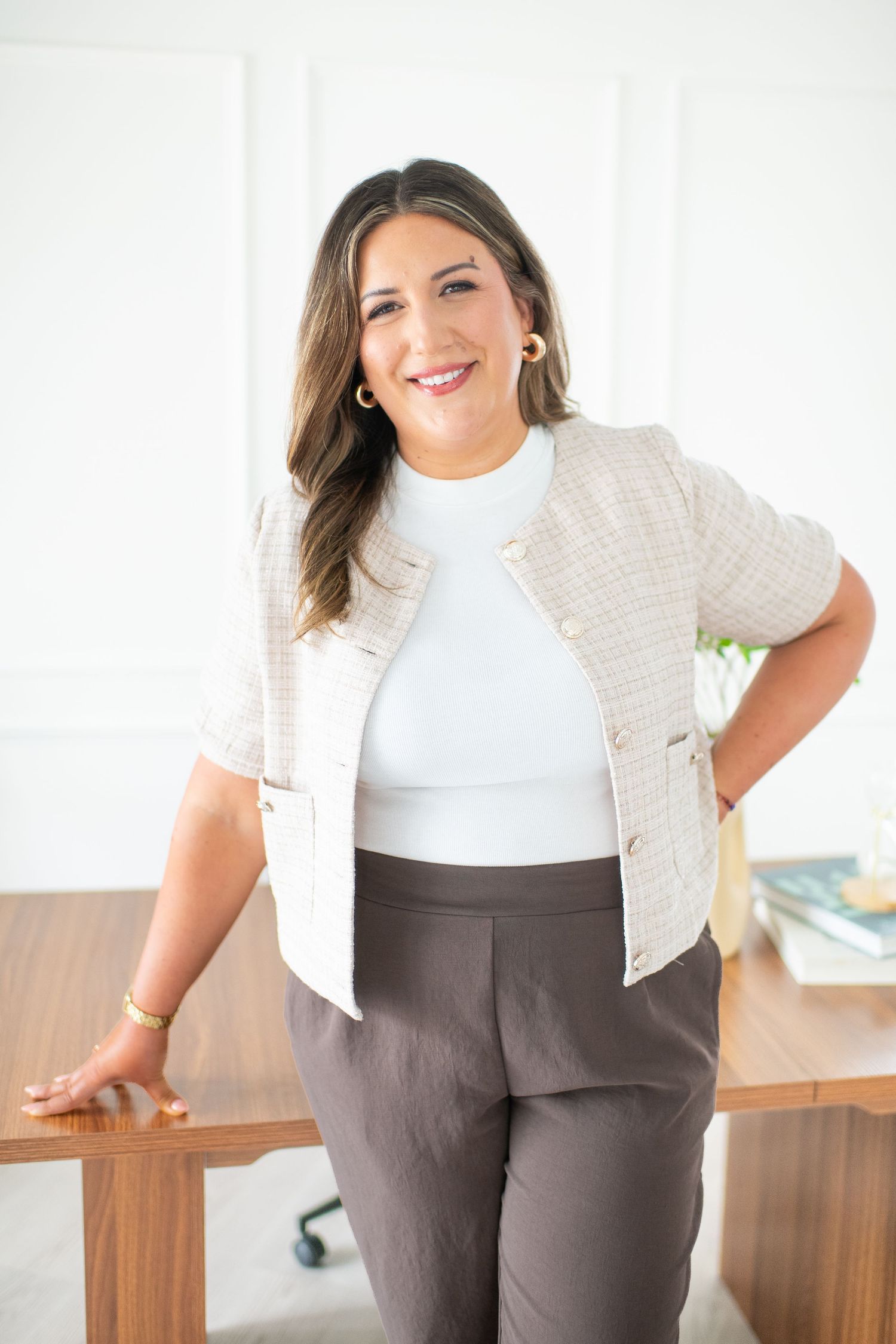 Professional woman in tweed blazer and gray trousers smiling confidently in a bright modern office setting.