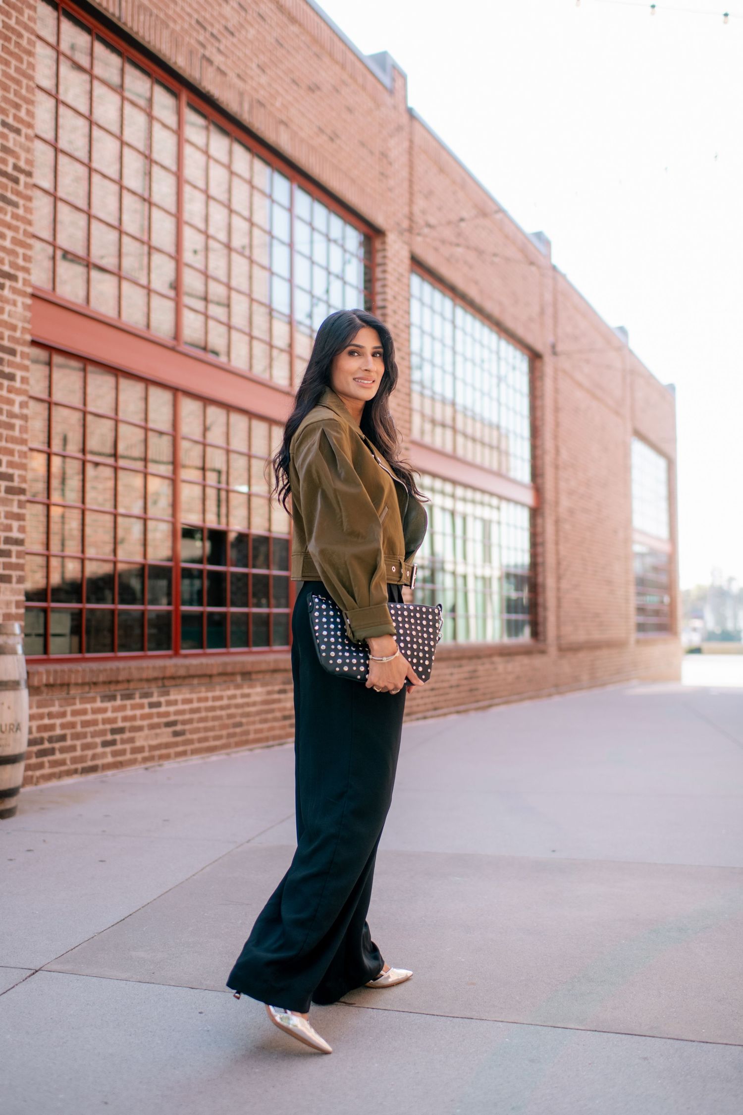 A person in a black dress and olive scarf poses in front of a brick building with large windows.