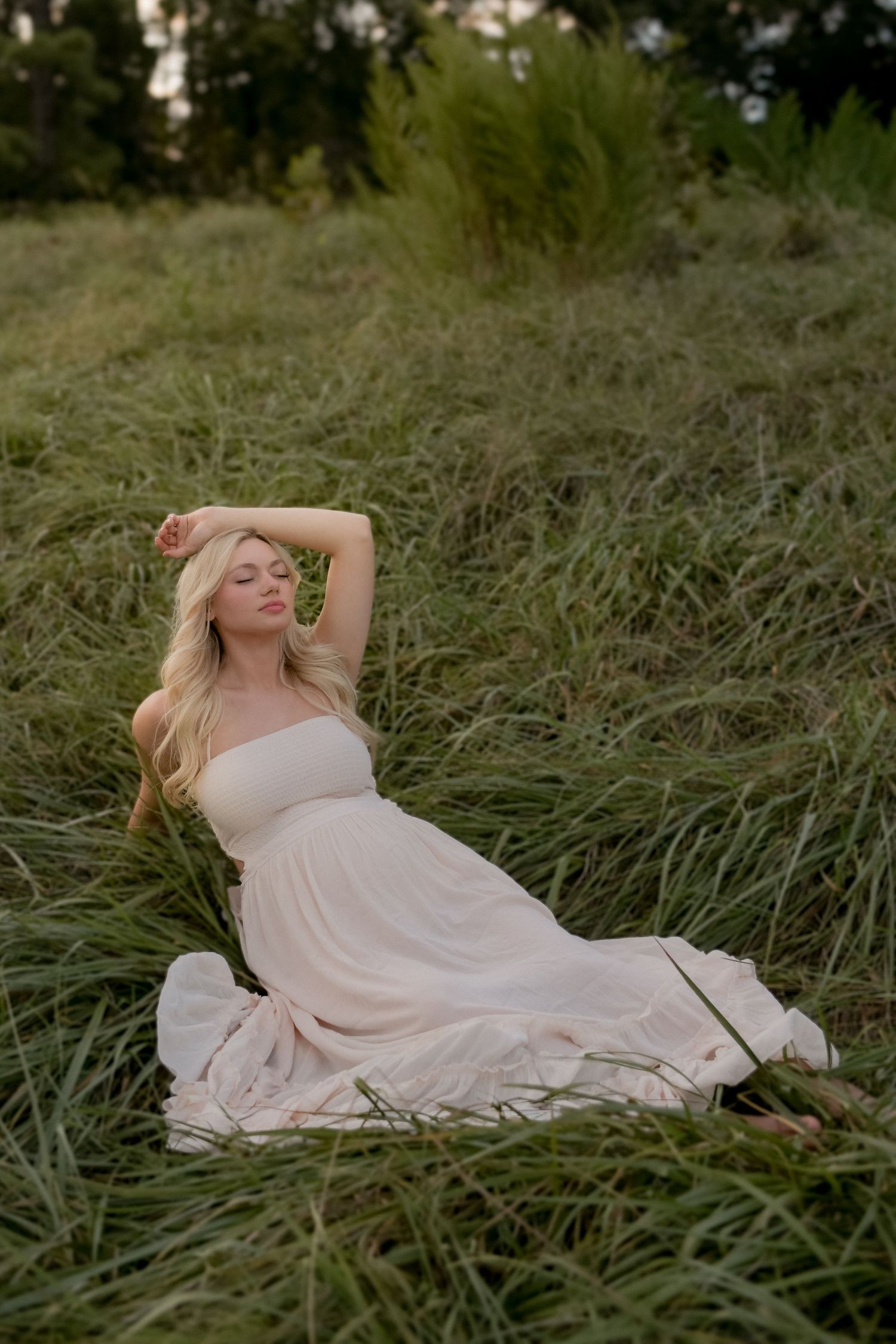 A woman in a flowy dress reclines gracefully in tall grass during a maternity photoshoot.