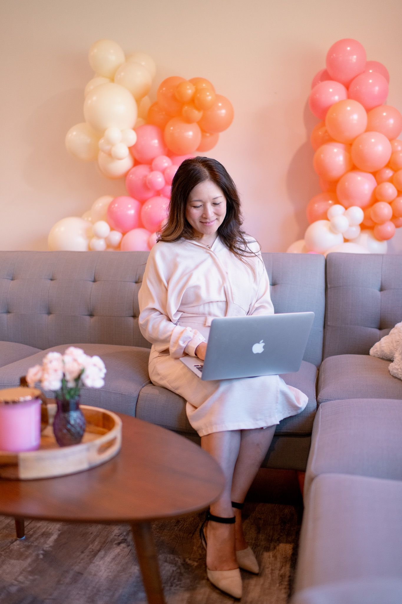 Person relaxing on gray couch with laptop while surrounded by pink and peach balloons in cozy indoor setting.
