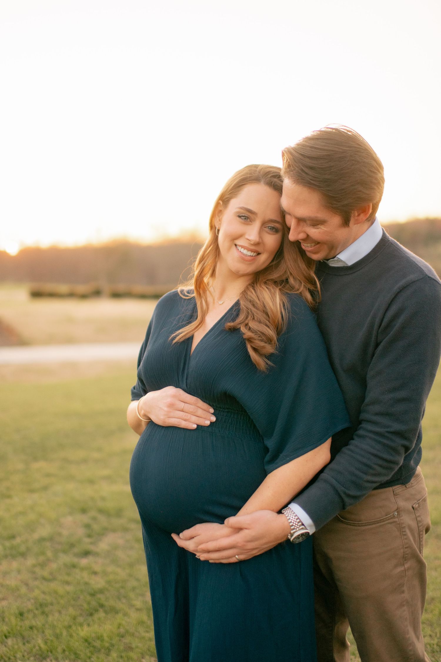 A loving couple embraces during sunset while cradling a baby bump, wearing matching navy outfits in a grassy field.
