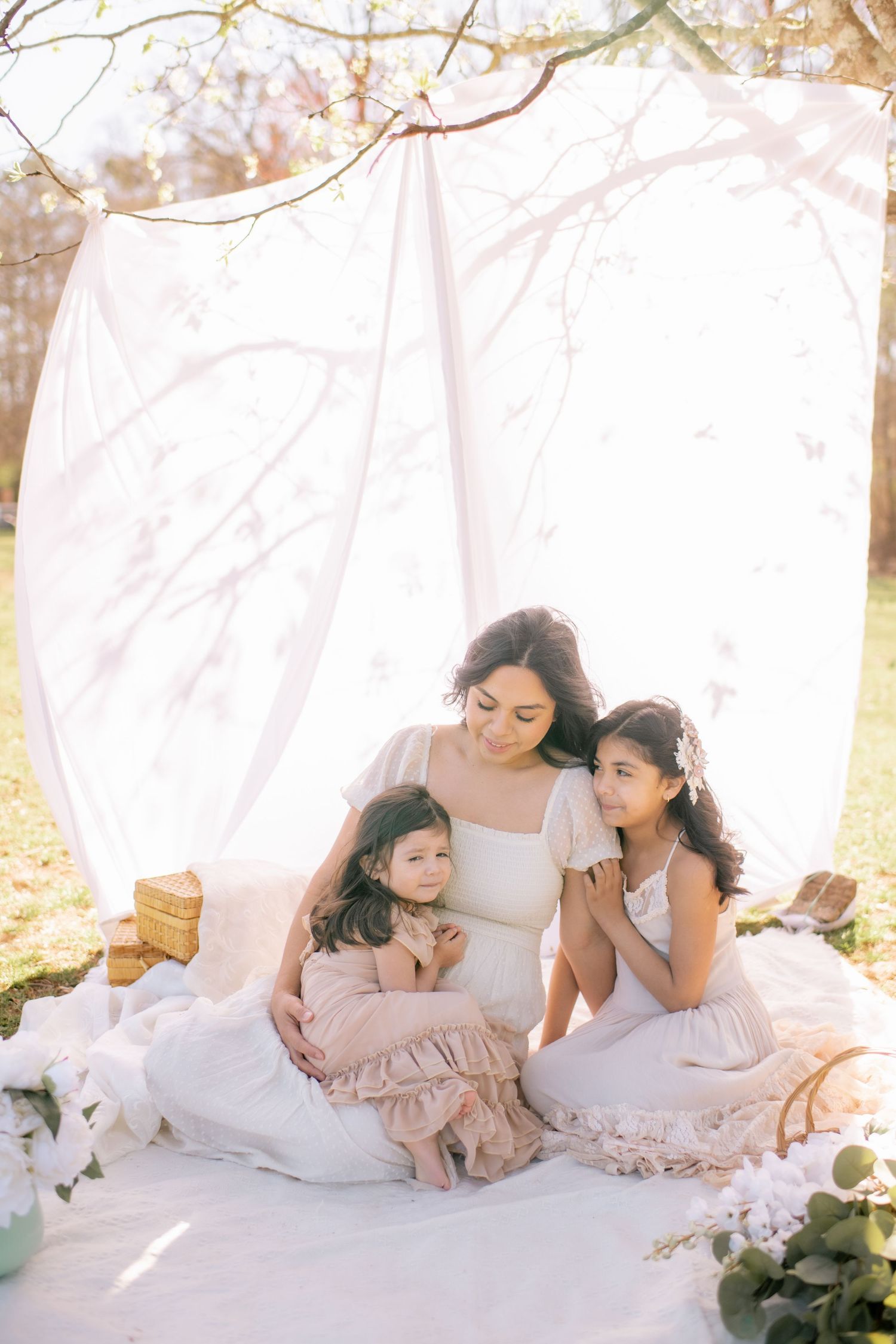 A soft and dreamy outdoor portrait scene with white flowing fabric and floral decor set in natural sunlight.