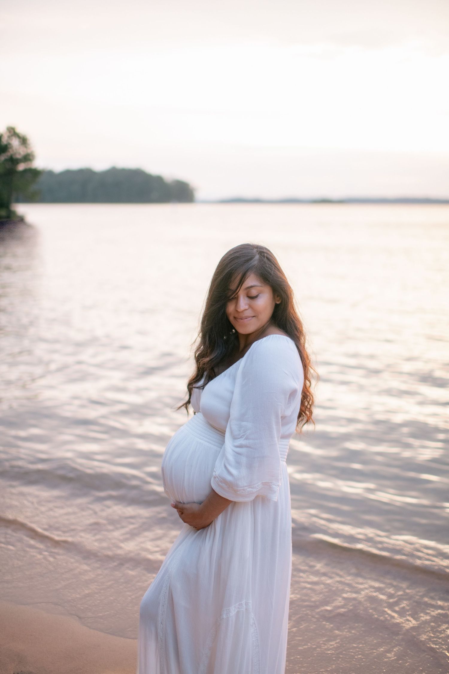 Expectant mother in flowing white dress poses on beach at sunset with lake and trees in background.