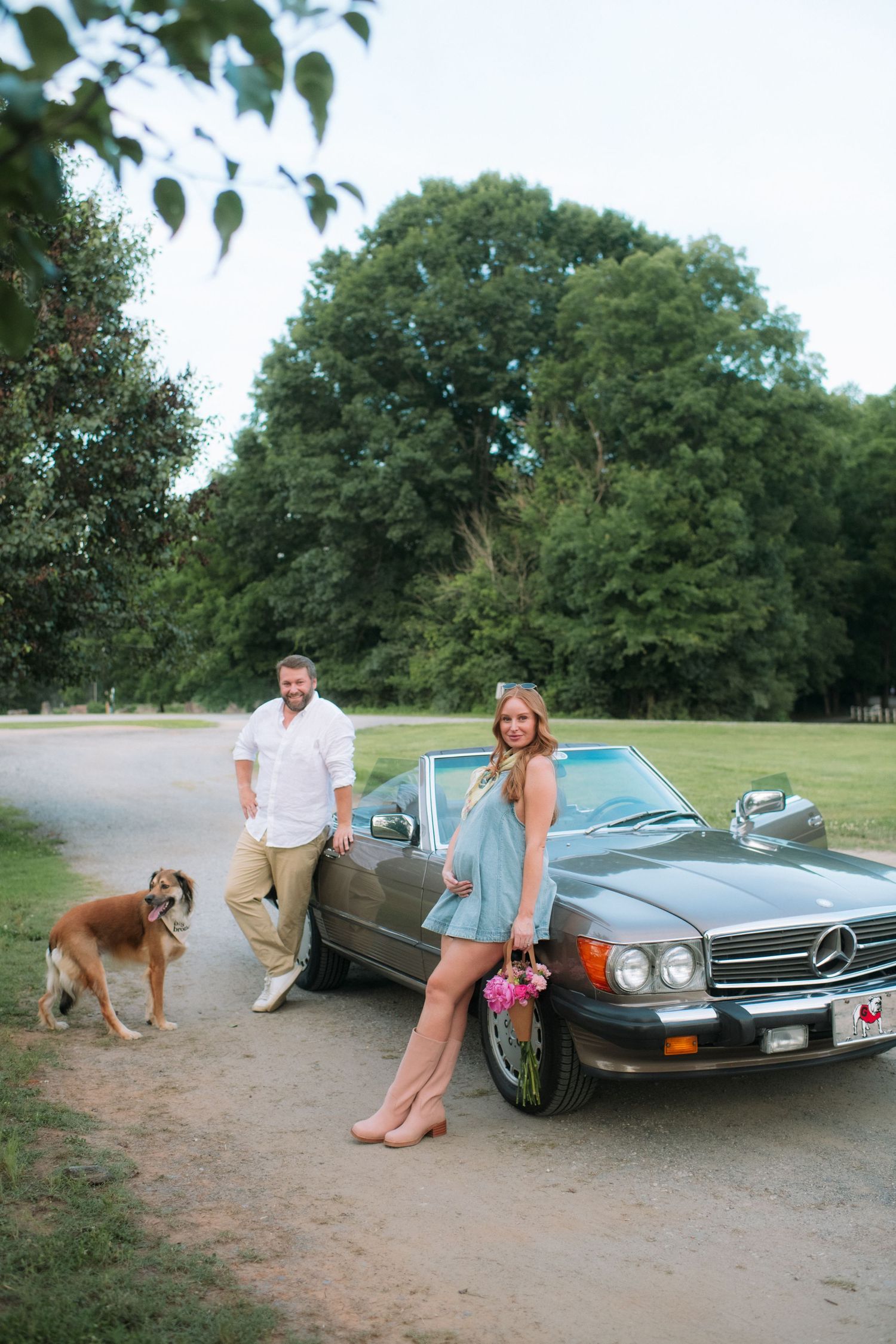 Couple posing with a classic Mercedes-Benz convertible and dog on a gravel path surrounded by lush green trees.
