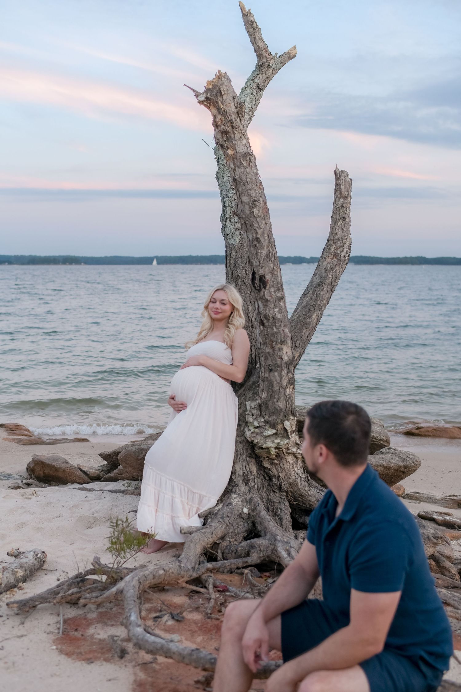 A couple shares intimate moments near a weathered driftwood tree on a lakeside beach.