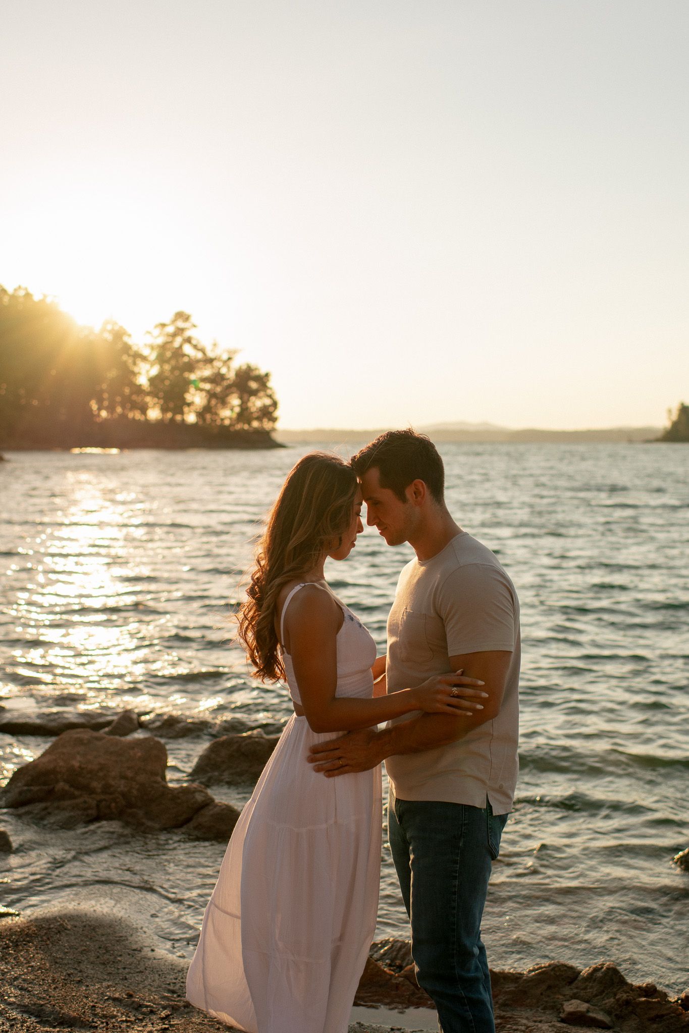 Romantic couple embraces at sunset by the water on rocky shoreline with golden sunlight streaming through trees.