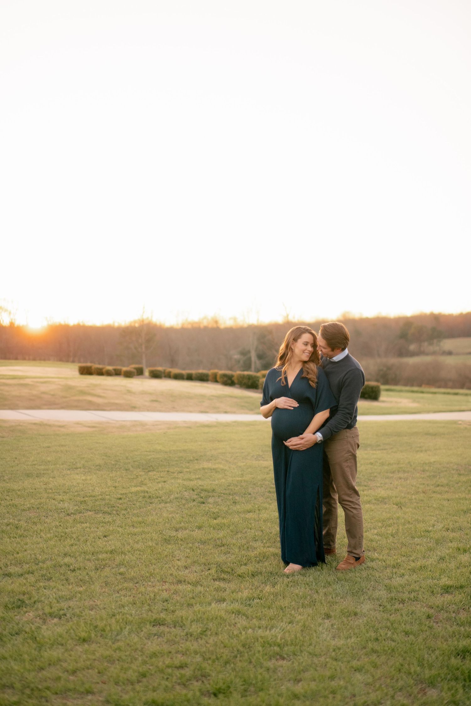 A couple embraces in a grassy field during golden hour sunset with rolling hills in the background.