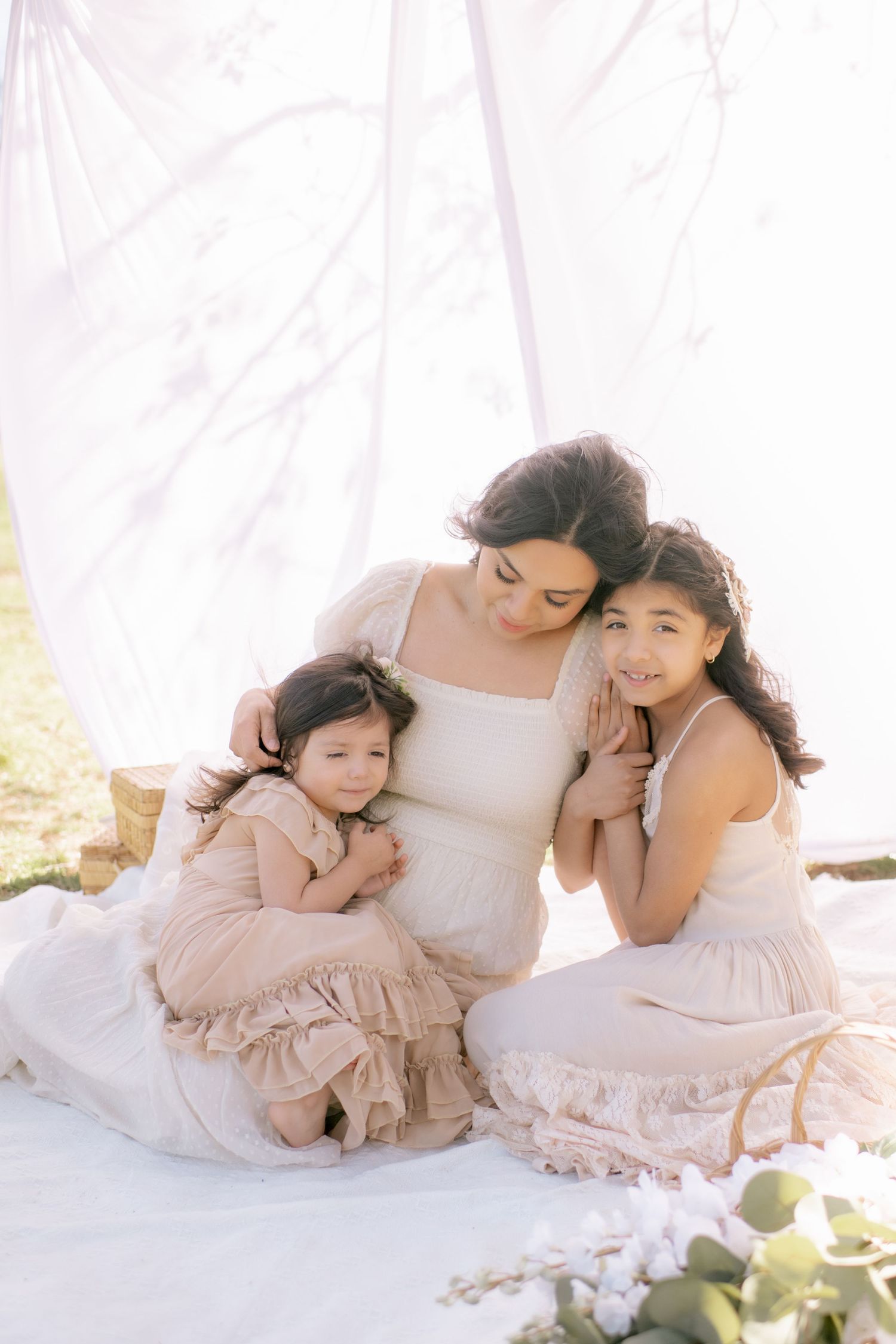 A heartwarming family portrait shows three people in white and beige dresses sitting close together on a light background.