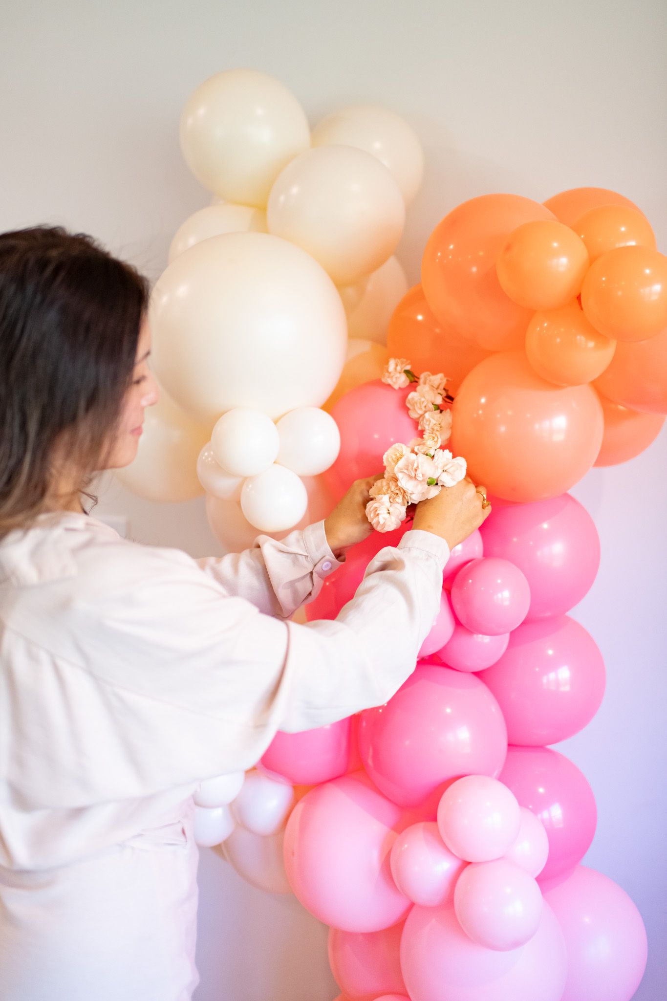 Person in white arranging a pink and peach balloon garland installation against light colored wall.