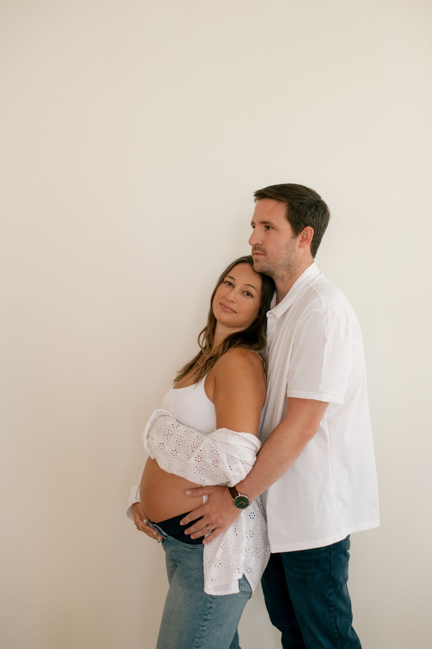 A couple embraces during an intimate indoor photoshoot against a white wall.