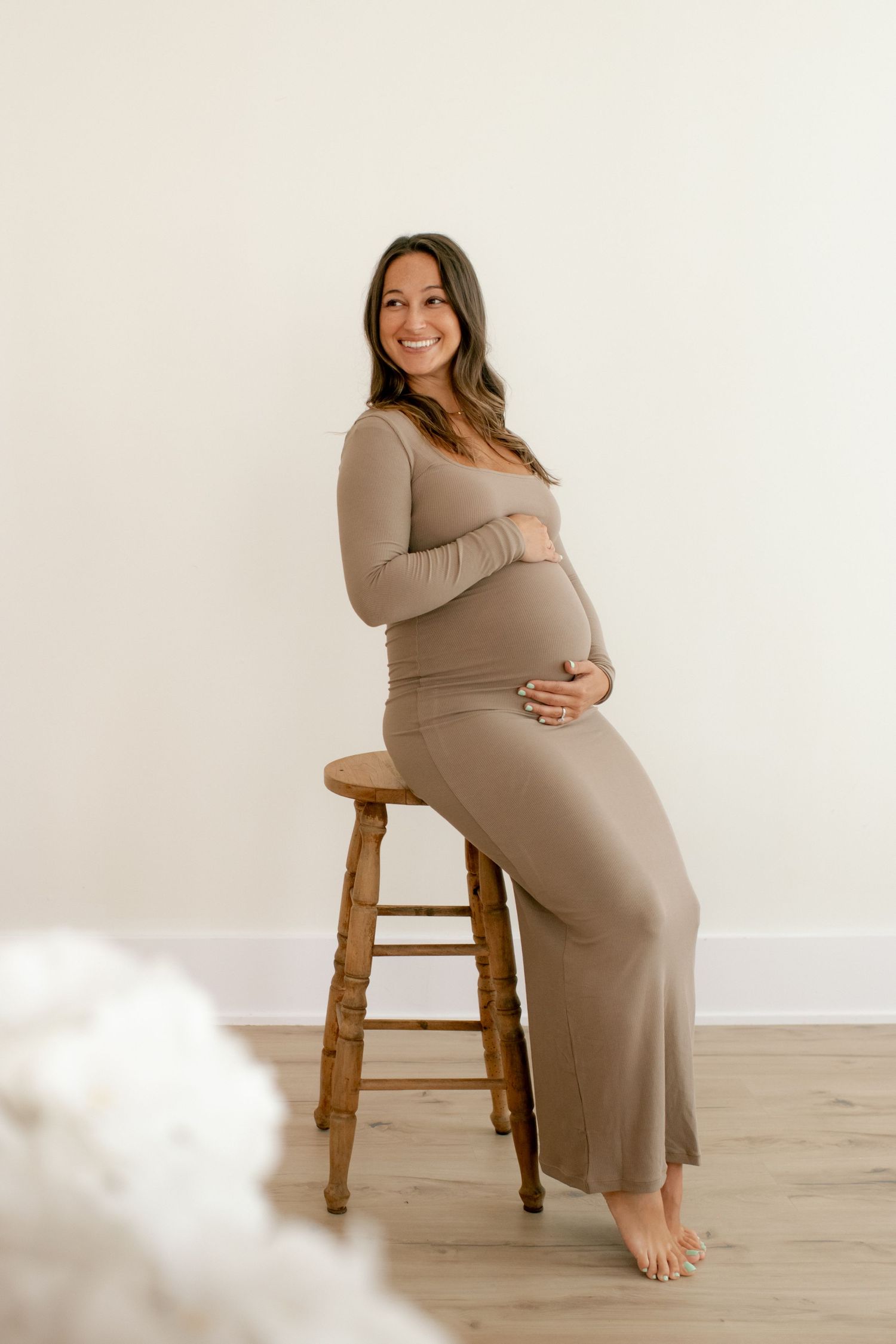 Pregnant woman in beige dress poses on wooden stool during maternity photoshoot.