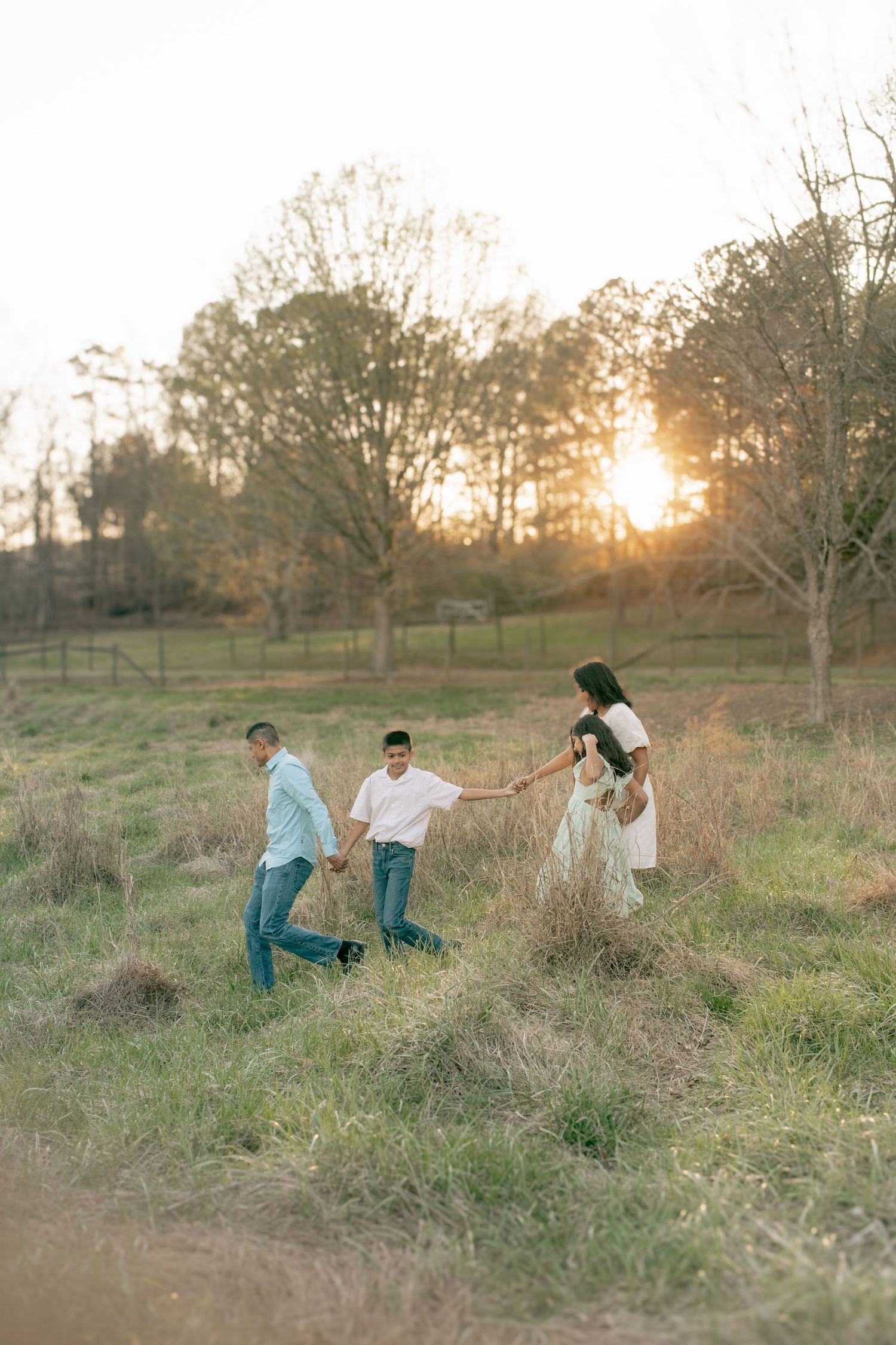 Three people holding hands and walking together in a grassy field at sunset with trees silhouetted in the background.