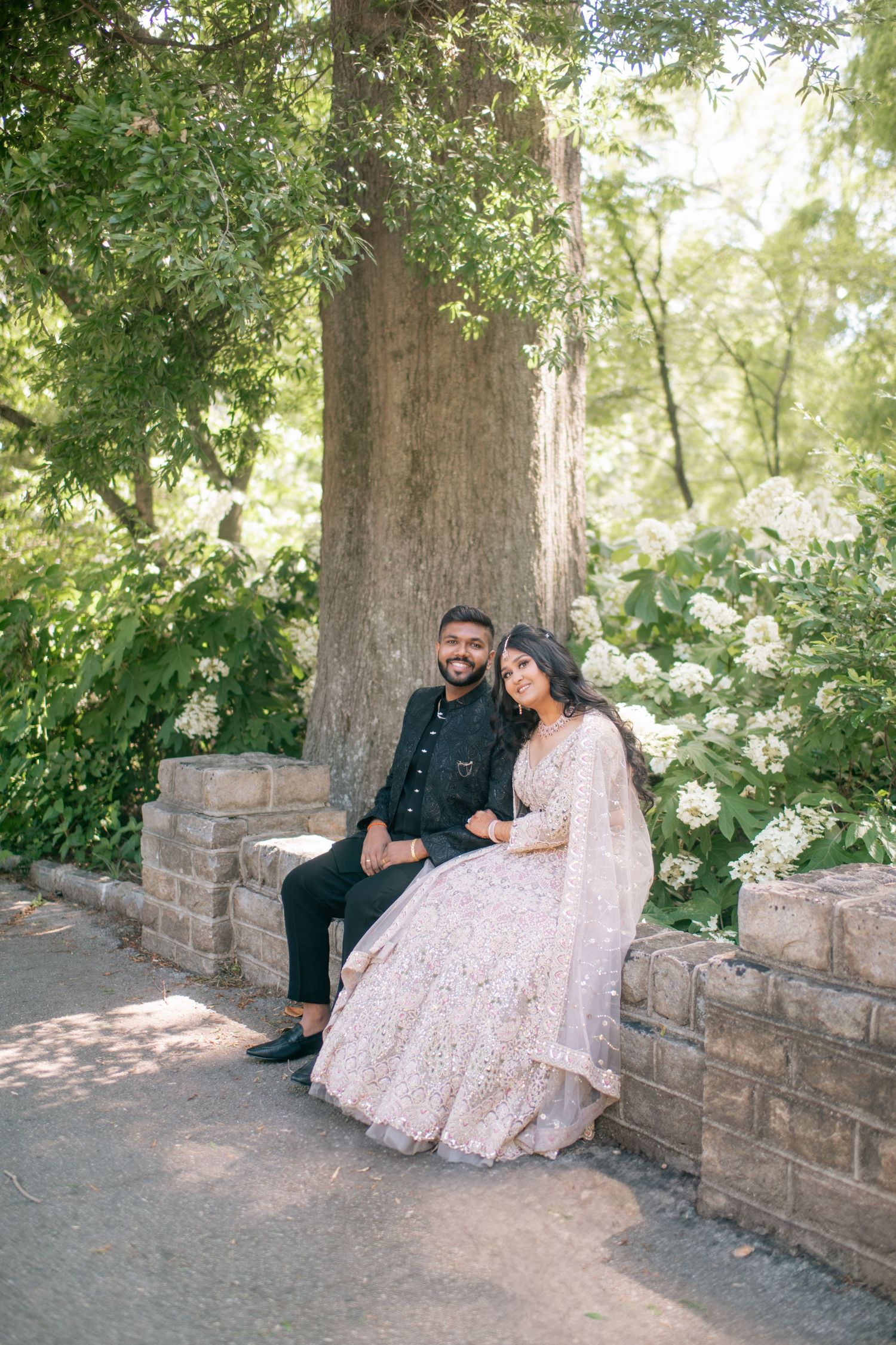 Two people share a romantic moment seated on stone wall under a large tree surrounded by blooming white flowers.