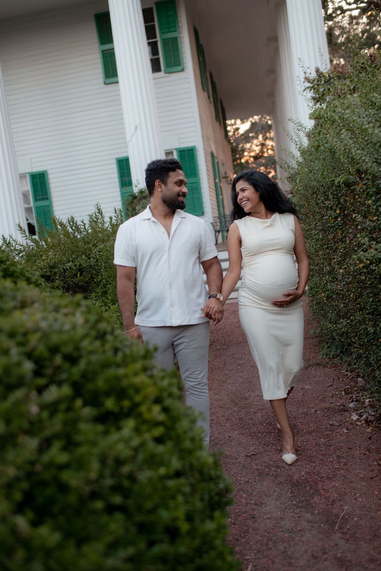 Couple in white outfits walking together on a garden path during a maternity photoshoot.