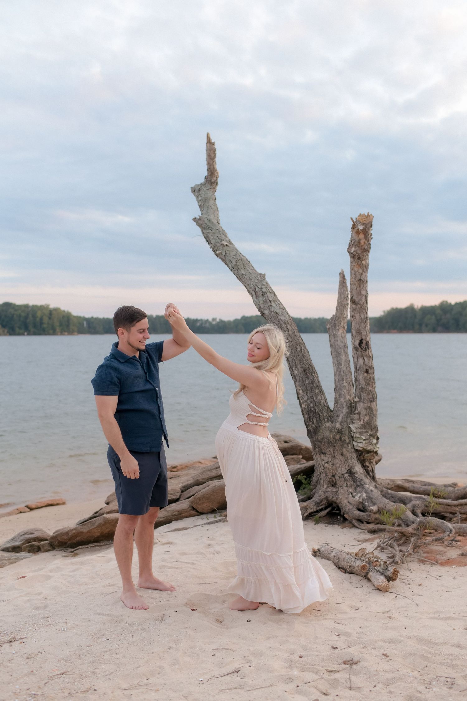 A couple dances playfully on a beach with driftwood and water in the background.