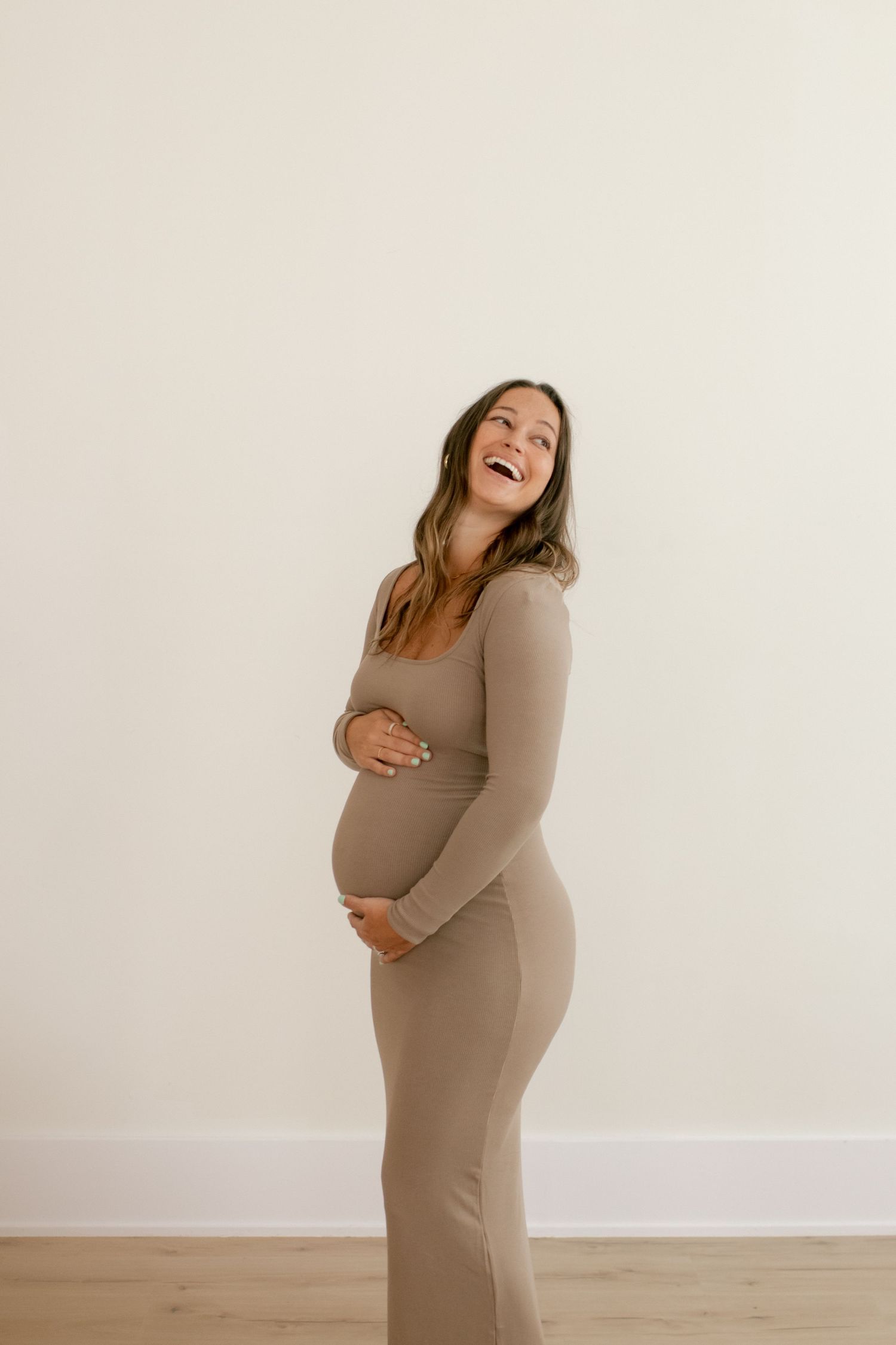 Joyful maternity portrait of woman in fitted beige dress against white wall.