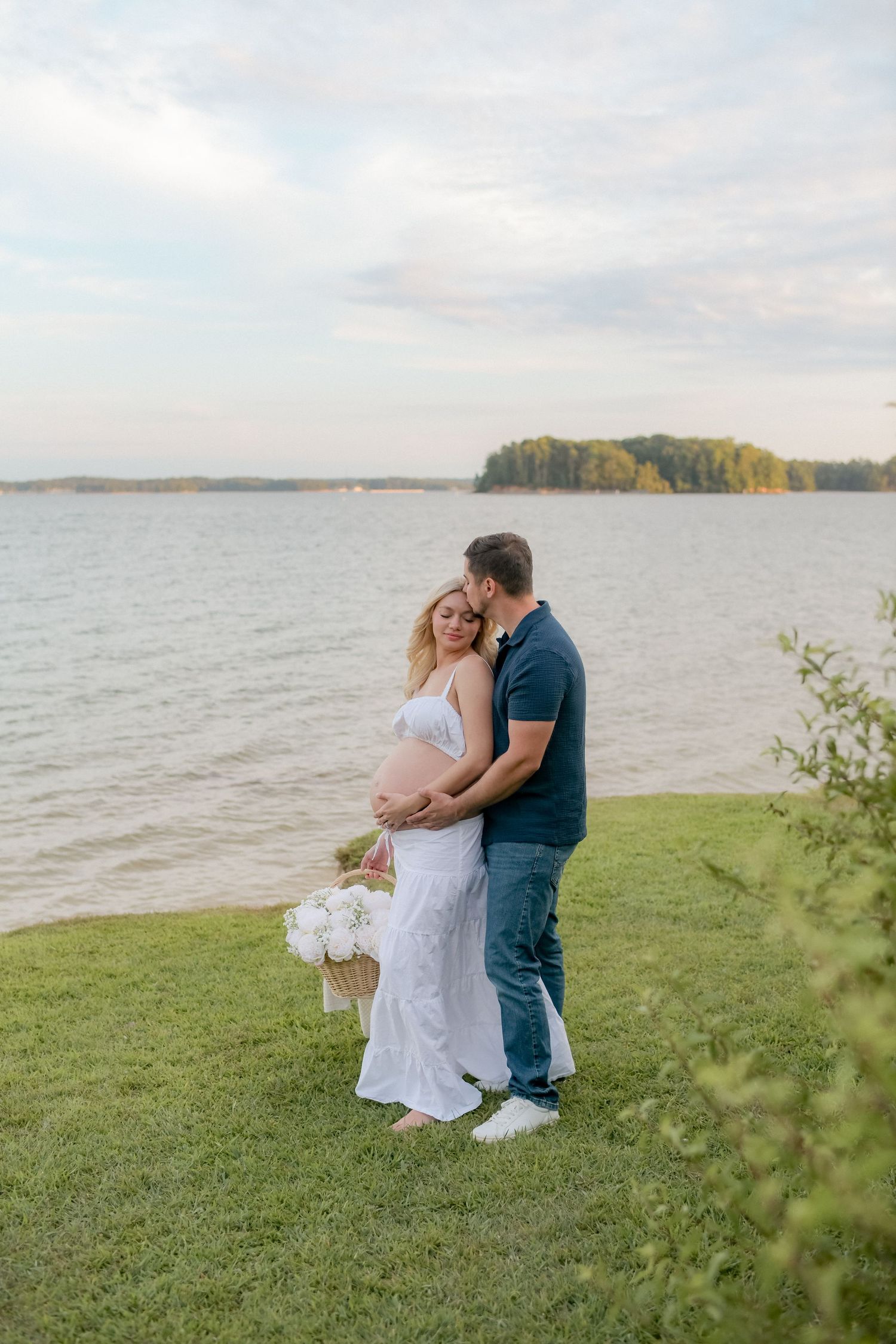 Waterfront maternity photos with couple embracing on grassy shore at sunset.