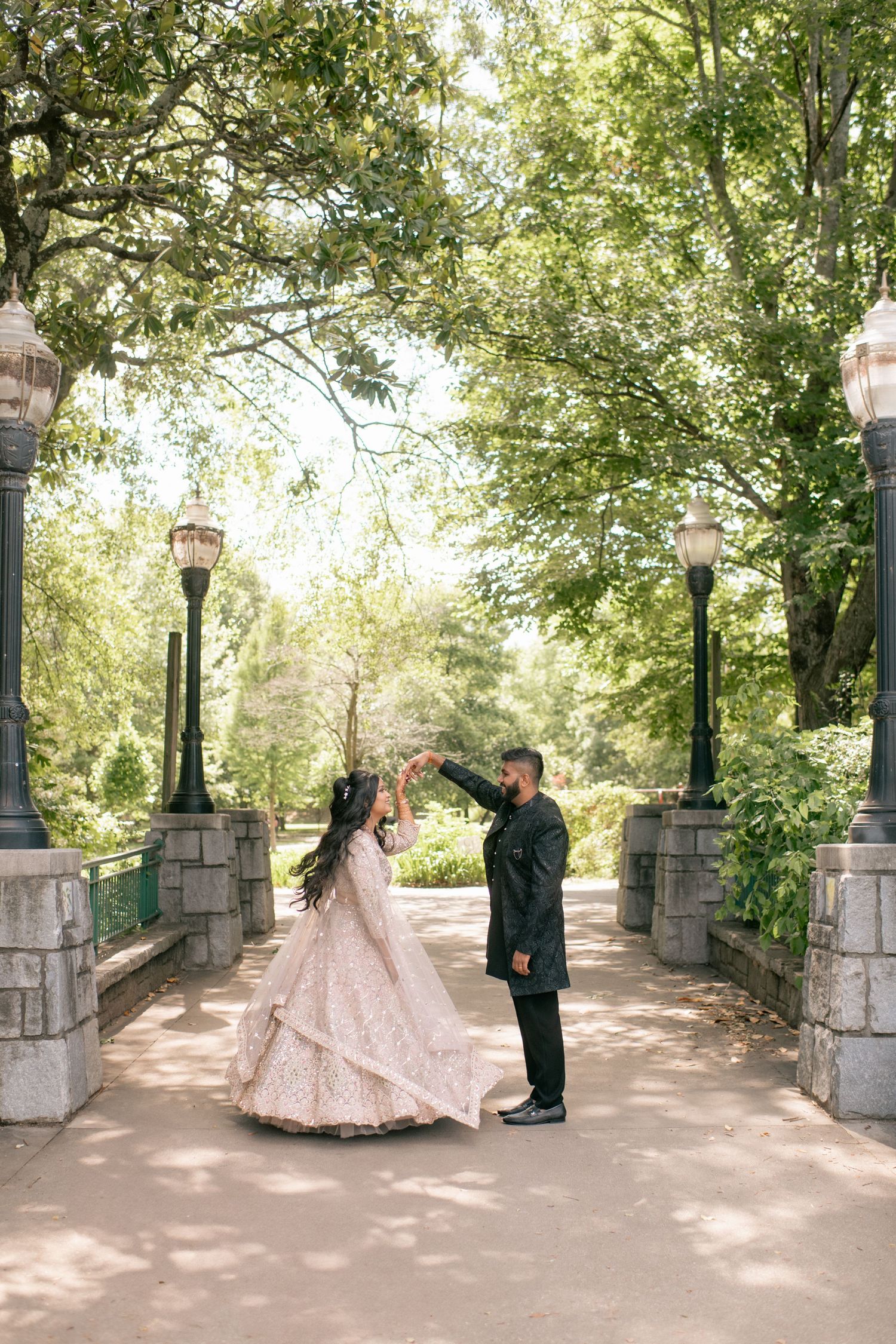 A couple in wedding attire poses along a tree-lined stone pathway in a romantic outdoor garden setting.