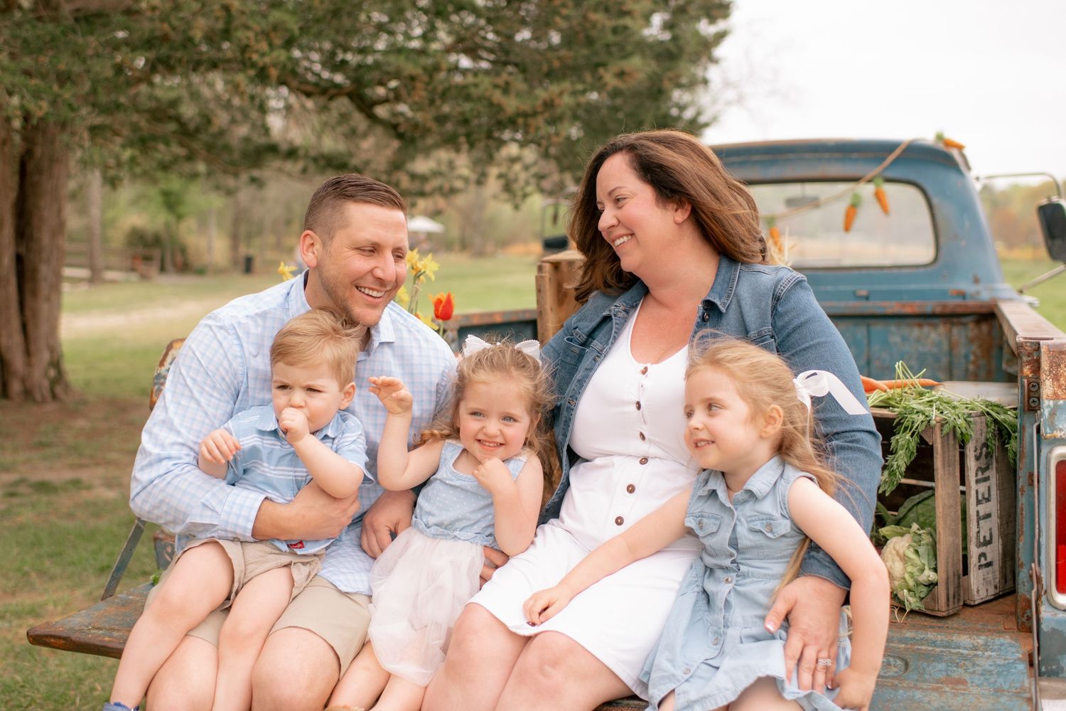 A family sits together on the tailgate of a vintage blue pickup truck in a rustic outdoor setting.