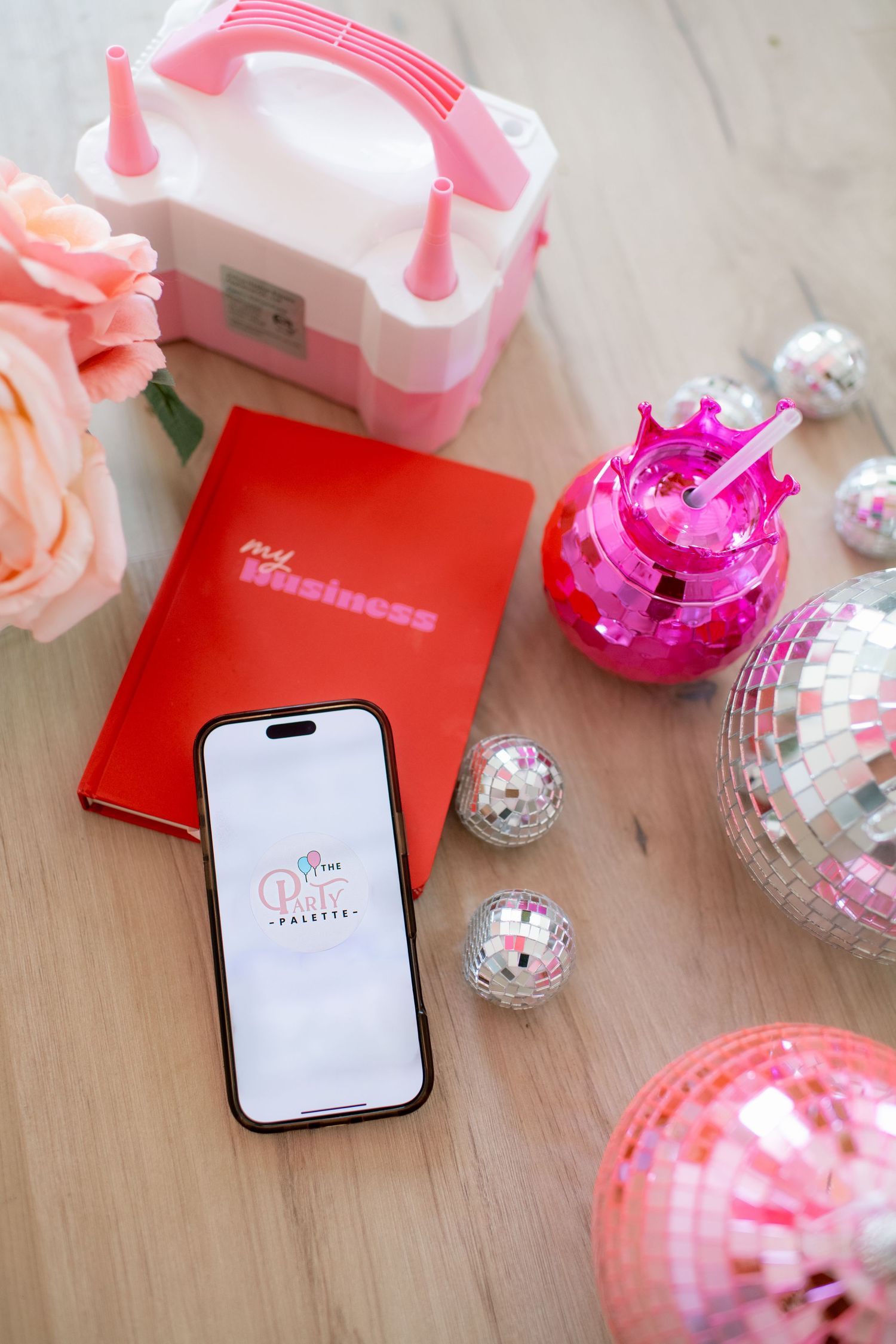 Phone display with pink accessories, disco balls and red notebooks arranged on wooden surface.