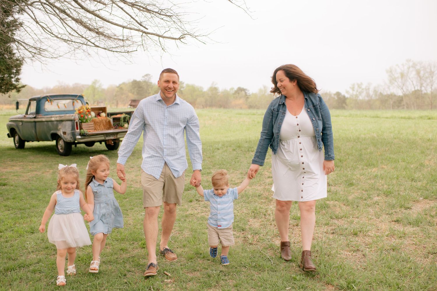 A family walks together through a grassy field on a sunny day with a vintage truck parked in the background.
