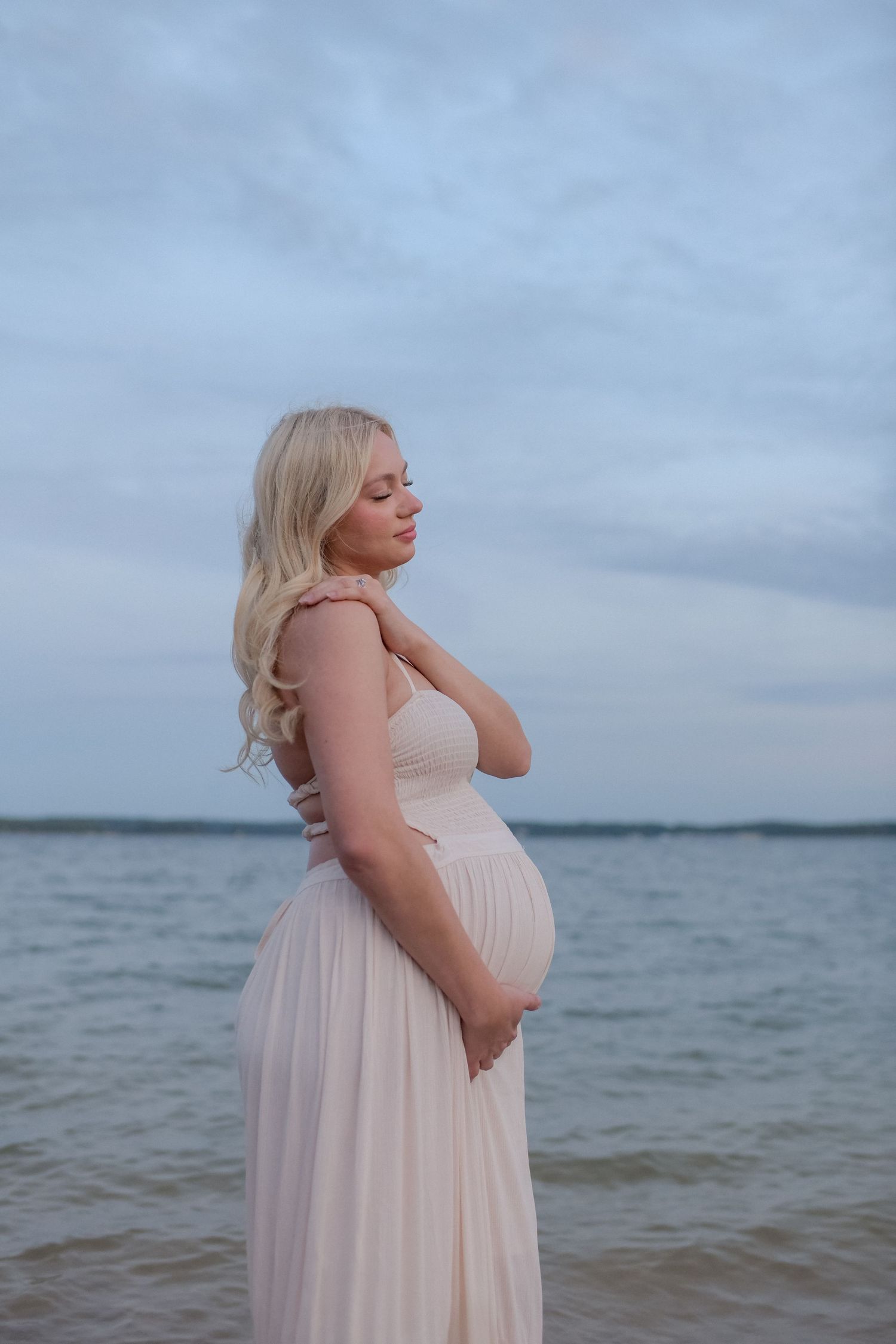 A woman in a flowing white dress poses by the ocean during sunset.