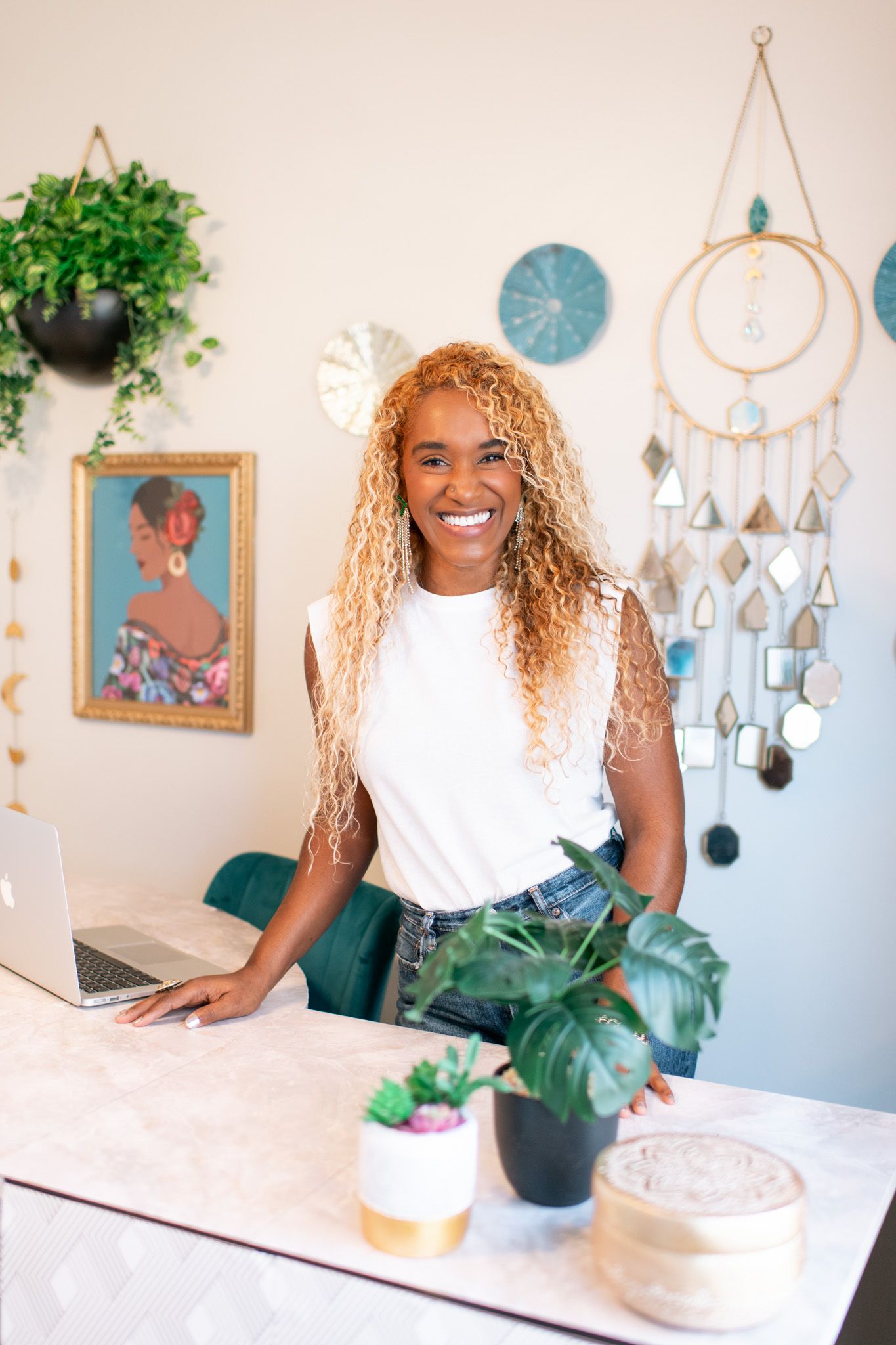 A person in a white shirt sits at a desk with plants and decorative wall hangings in a bright home office setting.