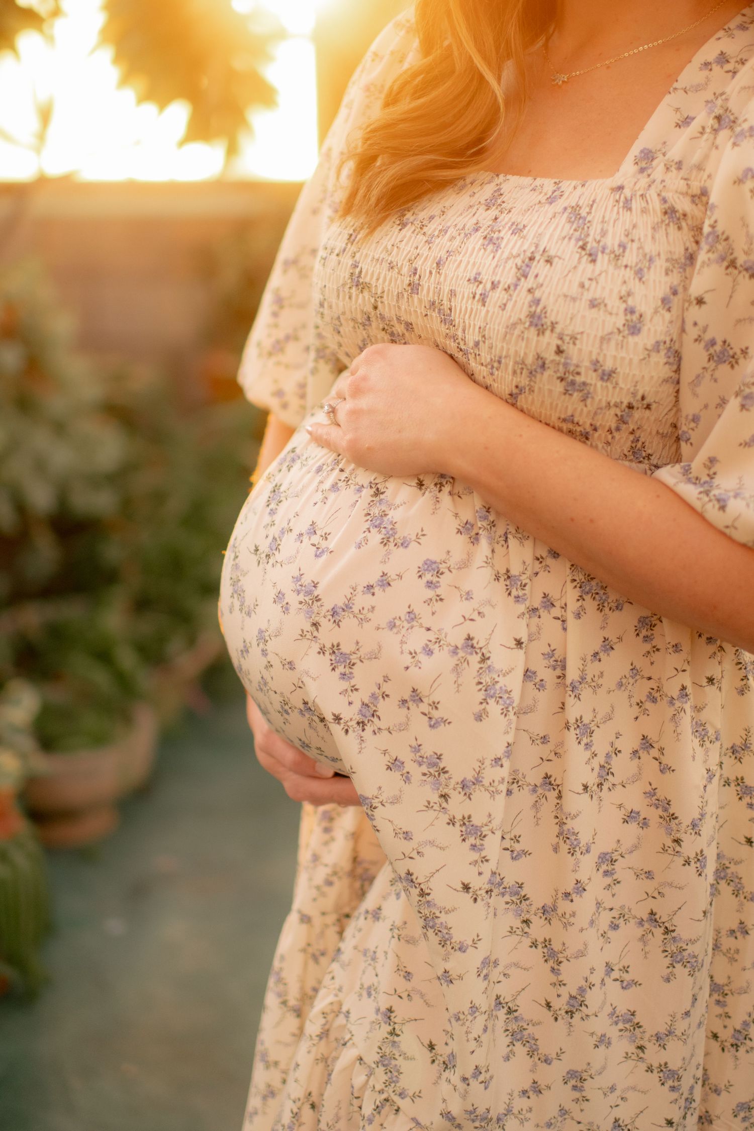 Close-up of a pregnant belly in a floral dress with golden sunset light streaming through a greenhouse window.